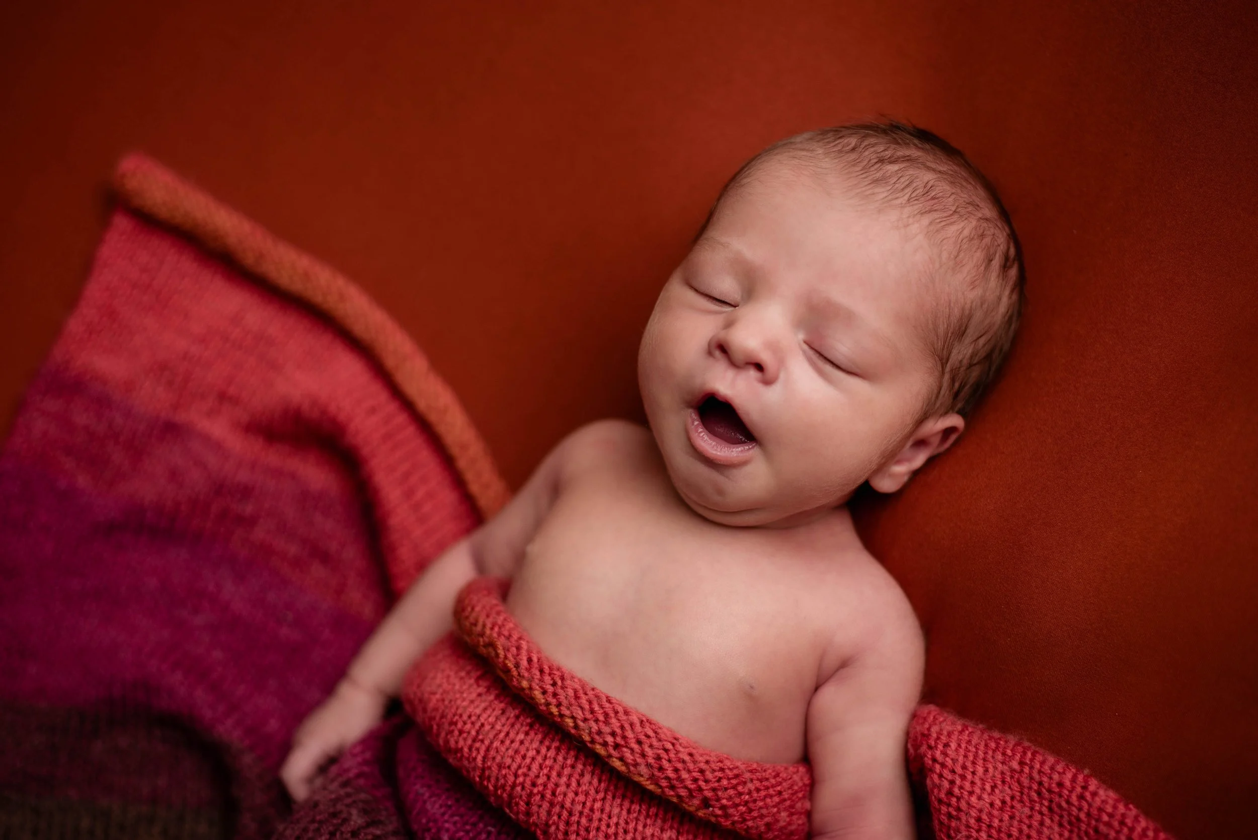 A sleeping newborn baby wrapped in a pinkish-red blanket with an orange rust background. Studio newborn portrait, Brownsville PA.