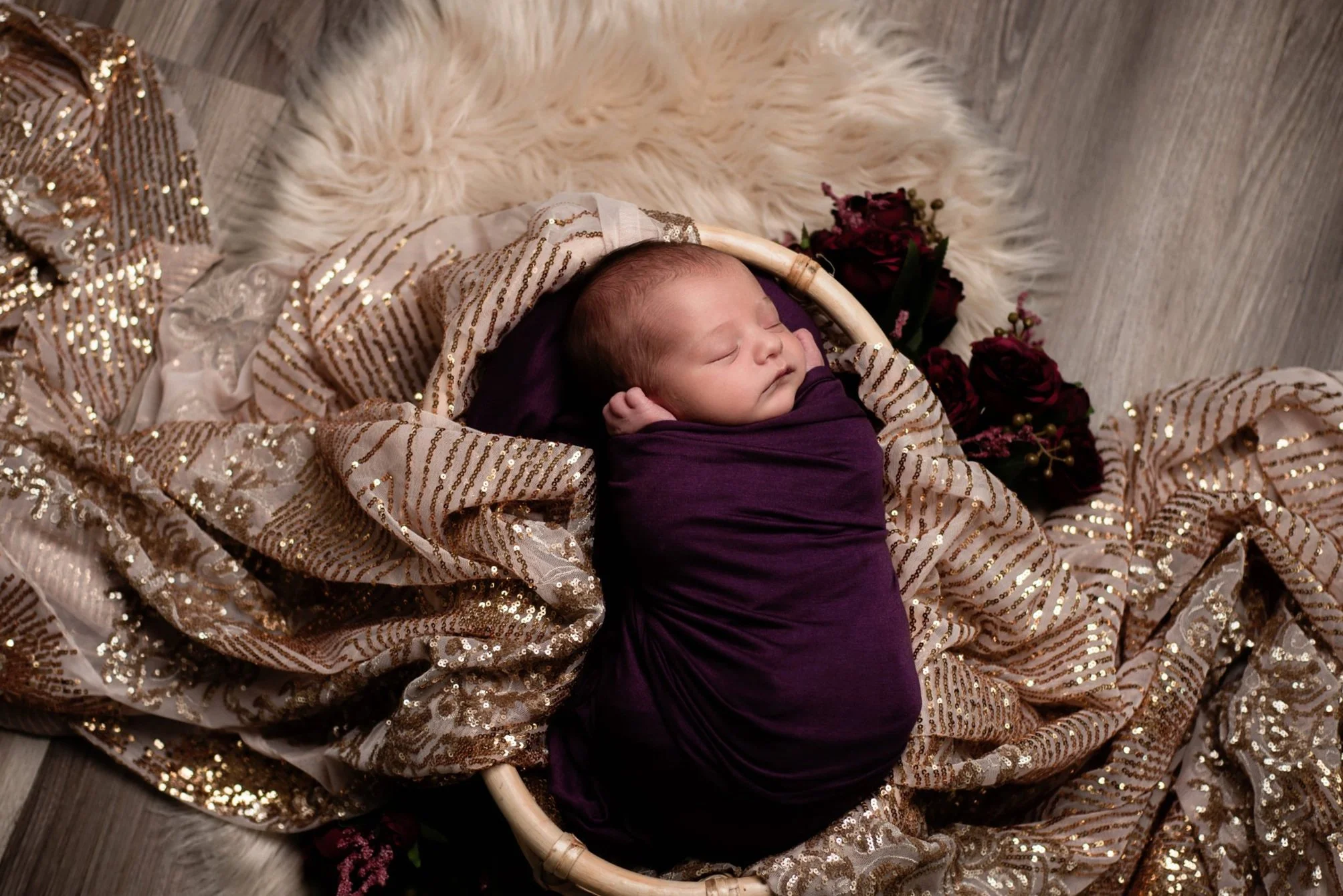 a one week old baby is swaddled in purple fabric, and lying in a basket draped with pink and gold sequined fabric, on top of a furry white blanket and deep red flowers