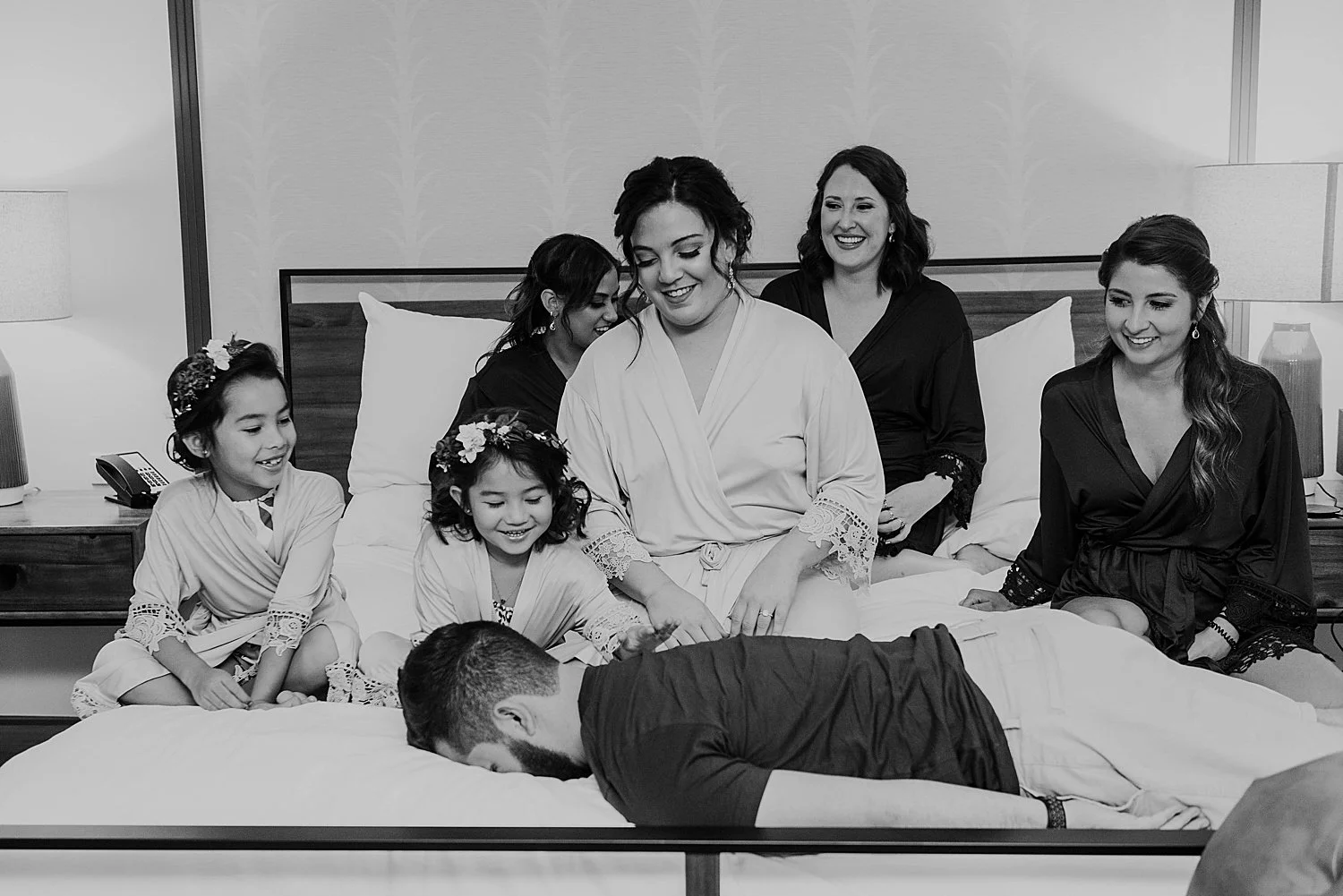 A man has face planted onto the bed, where a bride and her wedding party sit laughing.