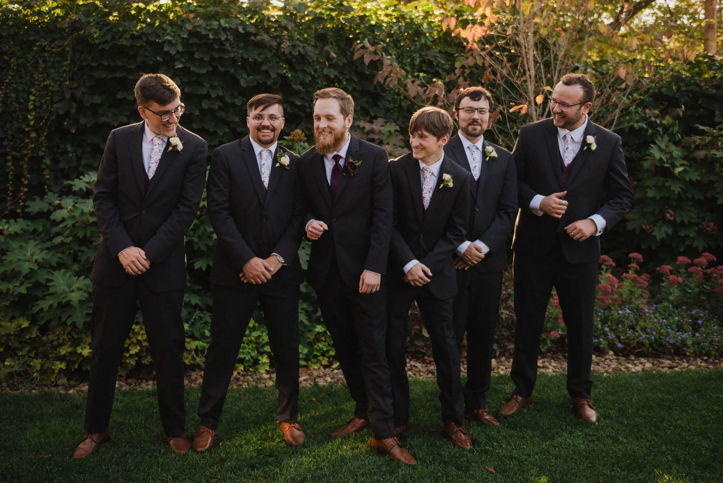 A group of seven men dressed in black suits, white shirts, and brown shoes, standing outdoors on grass with a backdrop of green and reddish-brown foliage. Groomsmen, smiling and laughing together. Wedding at Pittsburgh Aviary.
