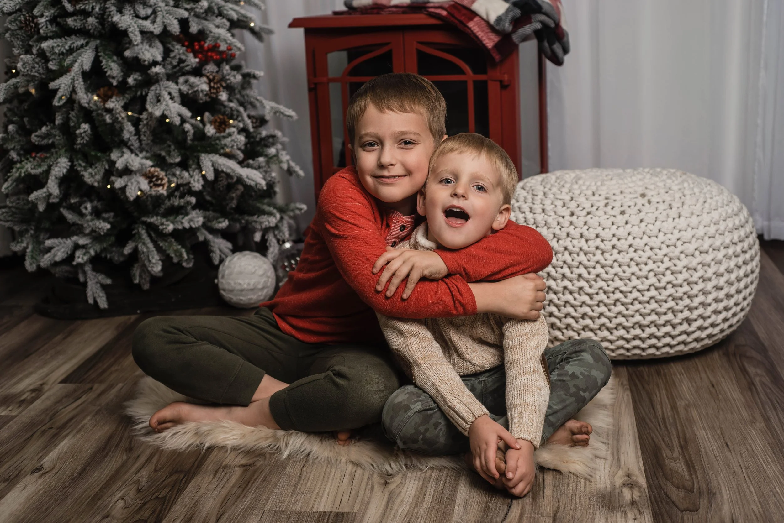 Two young boys hug each other while sitting on the floor on a white furry rug. Behind them is a red fireplace and a christmas tree.