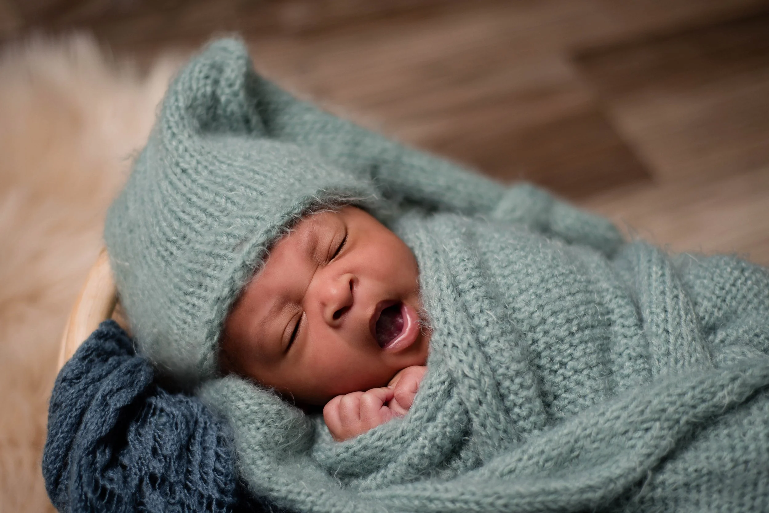 A newborn baby yawning, wrapped in a blue knitted blanket and wearing a matching hat, lying on a soft surface. Studio newborn portrait, Brownsville PA.