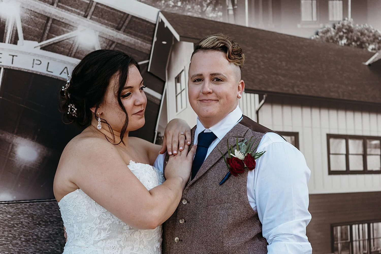 Groom looks at the camera smiling, he is a trans man with a nose ring wearing a brown tweed vest. Bride rests her hands on his shoulders and looks at his face, she is wearing a strapless lace gown.