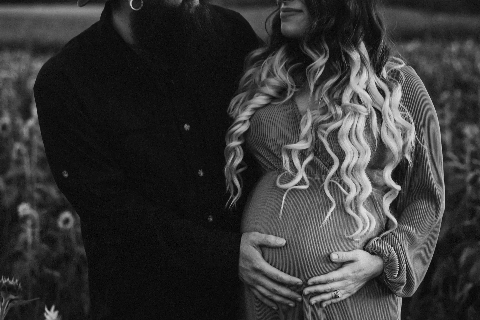 Black and white photo of a pregnant woman, she and her husband both have their hands on her belly, the top half of their faces aren't in view but she is smiling