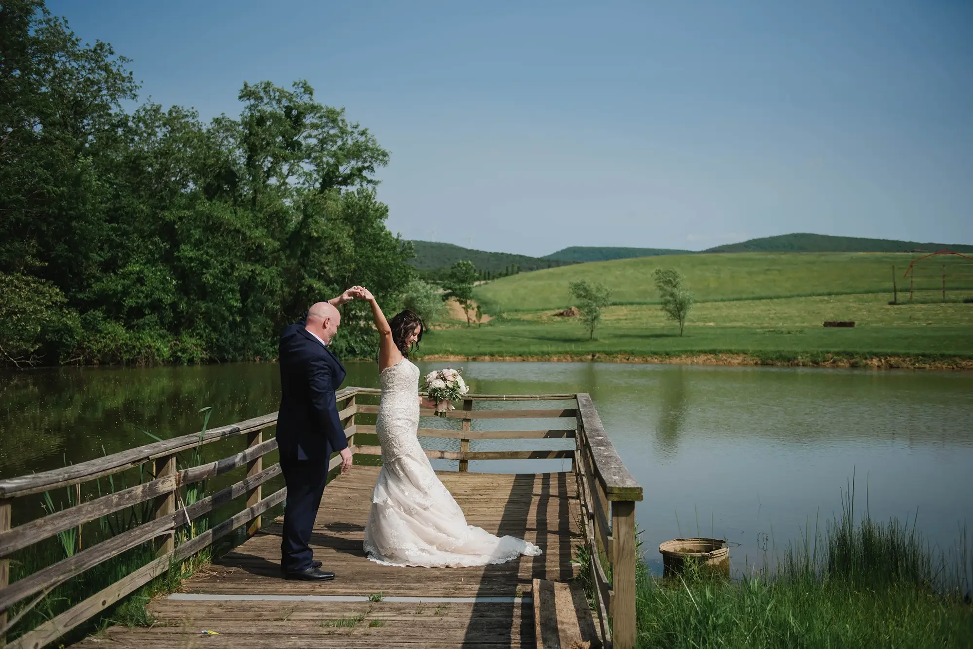 A bride and groom dancing on a wooden dock by a lake with green hills in the background on a sunny day. Microwedding at Rich's Farms.