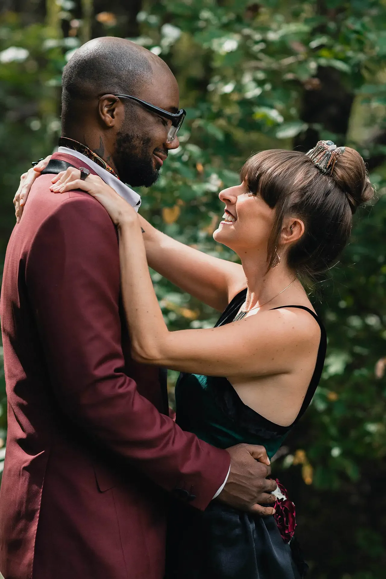 A black man in a maroon suit and a white woman with a high bun and a green jumpsuit smile at each other, they eloped at Raccon Creek State Park, and the background is all greenery
