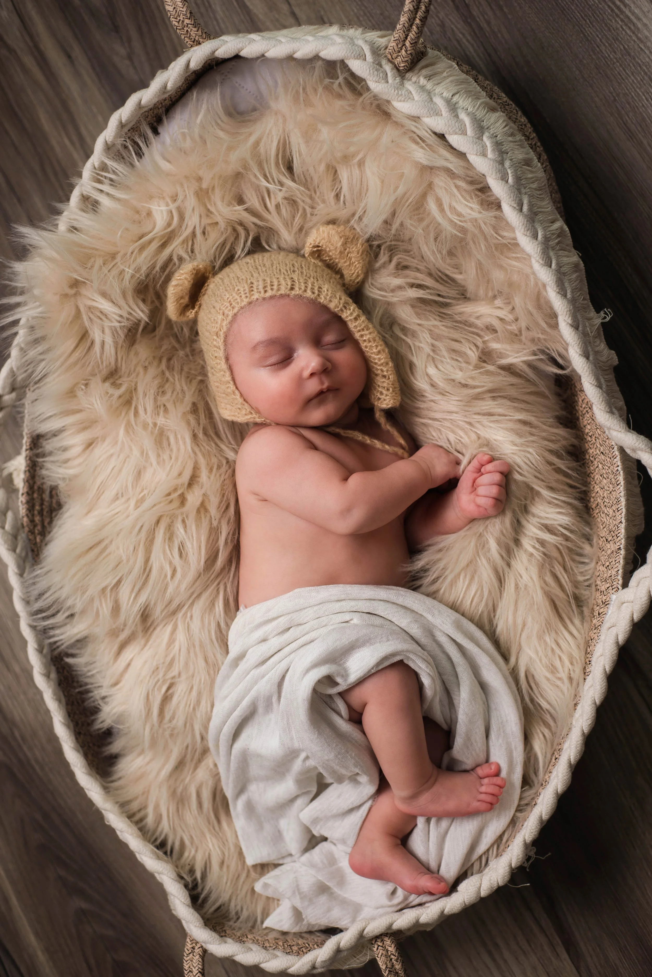 A sleeping baby wearing a beige bear hat, wrapped in a white cloth, lying on a fluffy cream-colored blanket in a woven basket. Studio newborn portrait, Uniontown PA.
