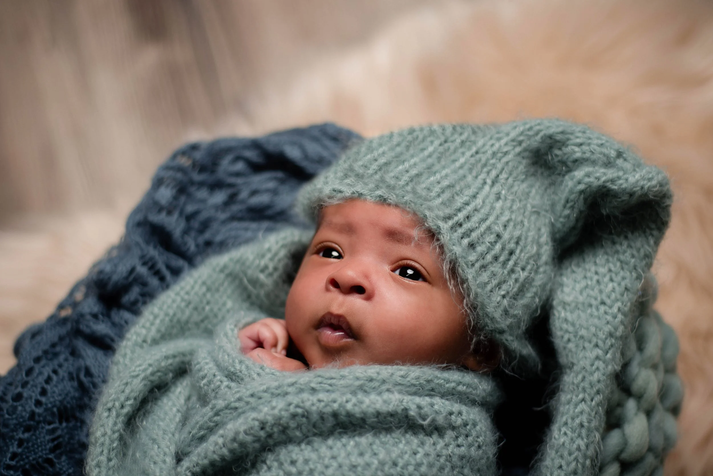 Close-up of a baby bundled in gray knitted blankets and wearing a blue knitted hat, lying on a soft surface. Studio newborn portrait, Uniontown PA.