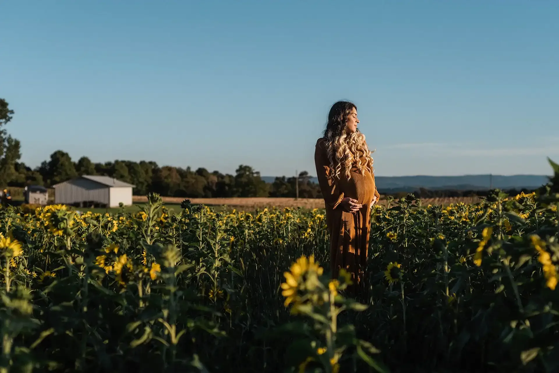 A pregnant woman stands in a sunflower field during golden hour, a peaceful expression on her face. she wears a long corduroy gown.