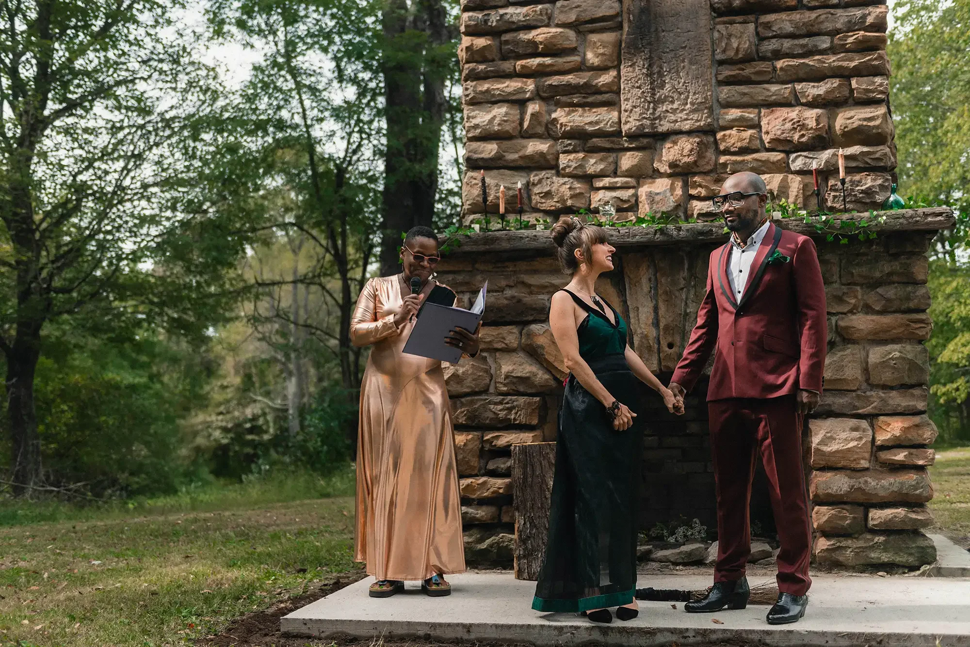 A couple holding hands during a wedding ceremony outdoors, with a woman officiant reading from a book nearby. Elopement in Racoon Creek State Park, PA