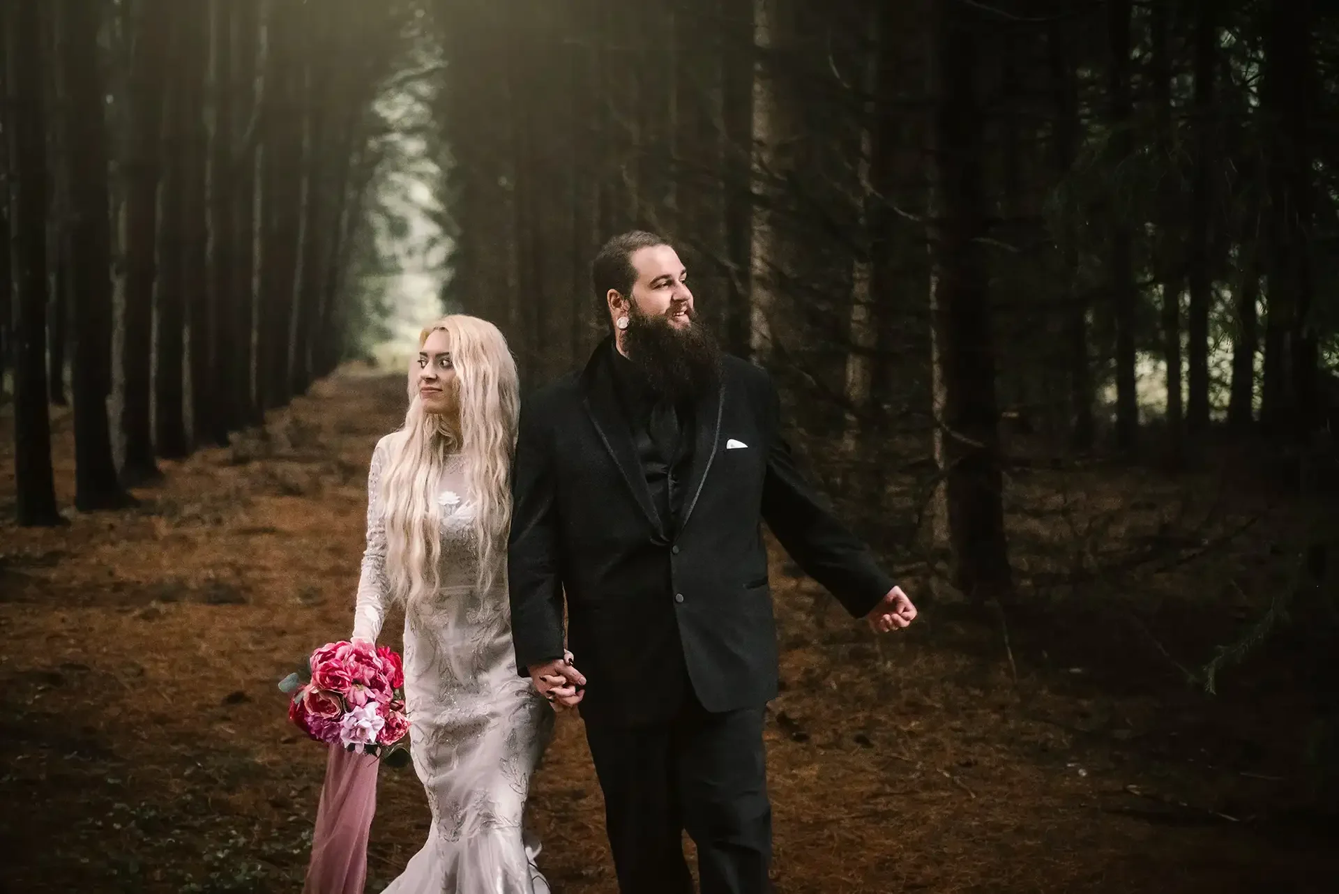 a bride and groom stand in a dark forest, they are facing away from each other but holding hands