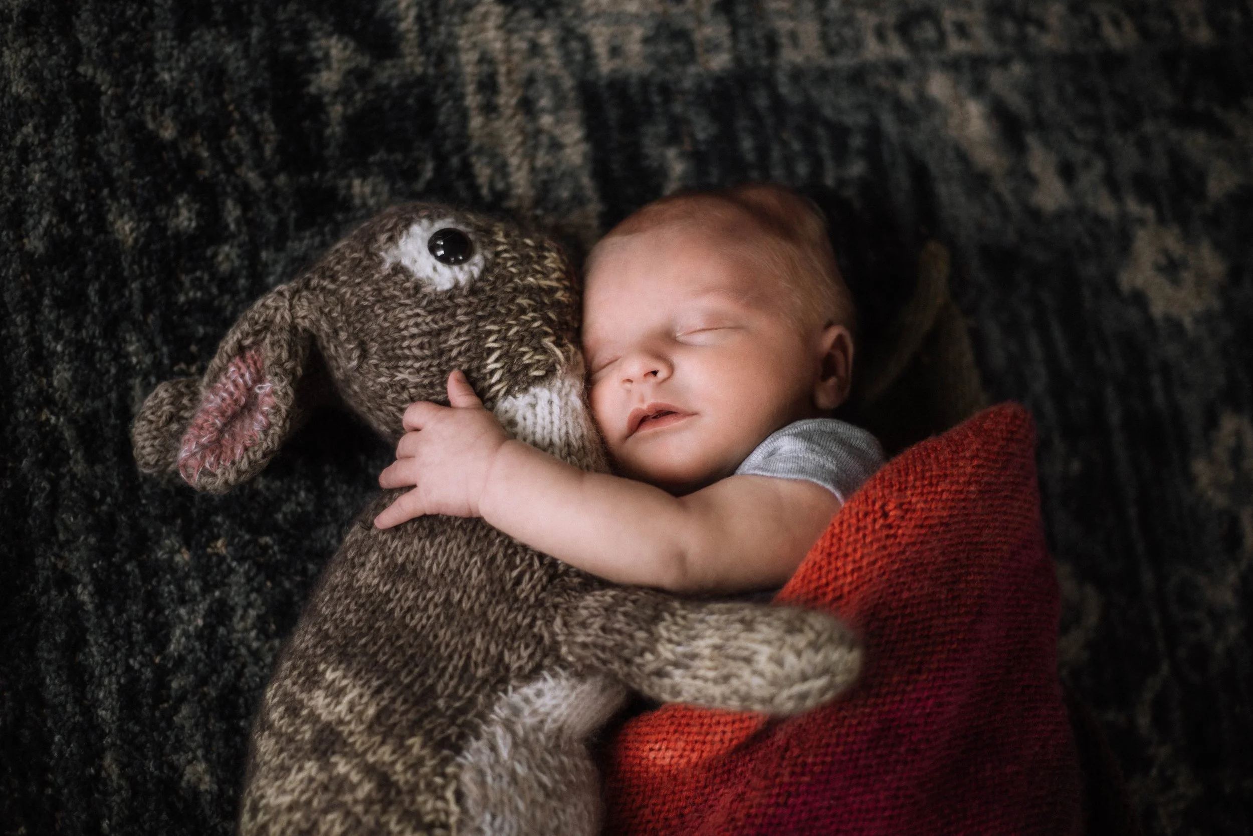 A sleeping baby hugging a knitted stuffed animal rabbit on a dark textured surface. Studio newborn portrait, Brownsville PA.