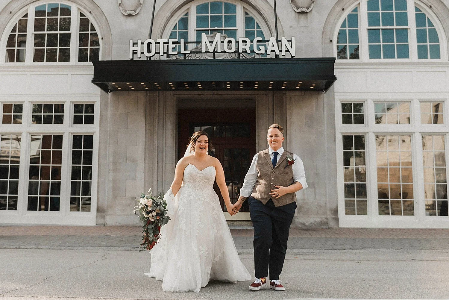 a trans man is dressed in a vest and dress pants, holding hands with his new wife dressed in a gown, in front of the Hotel Morgan.