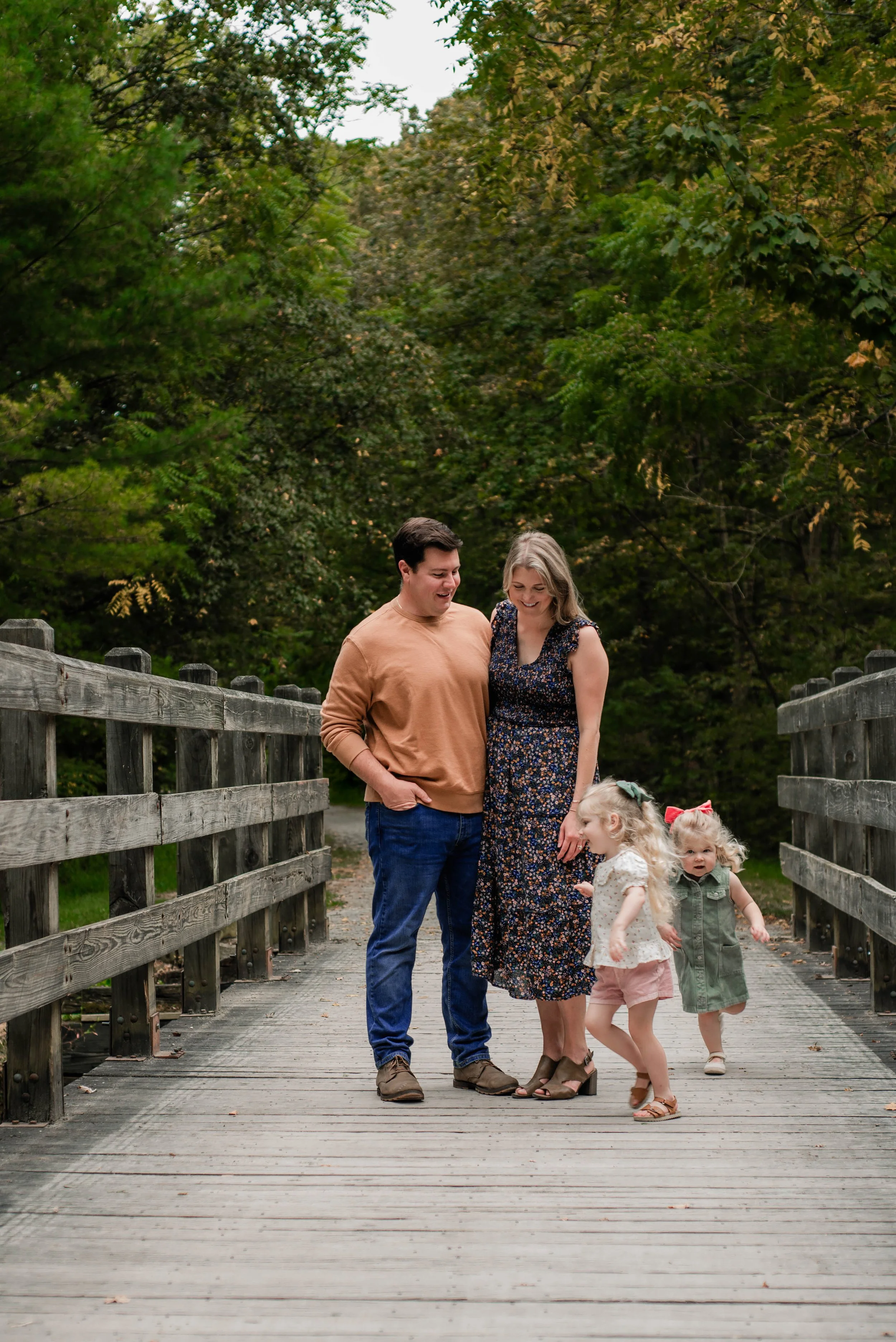 mom and dad stand on a wooden bridge while their young daughters run in circles around them