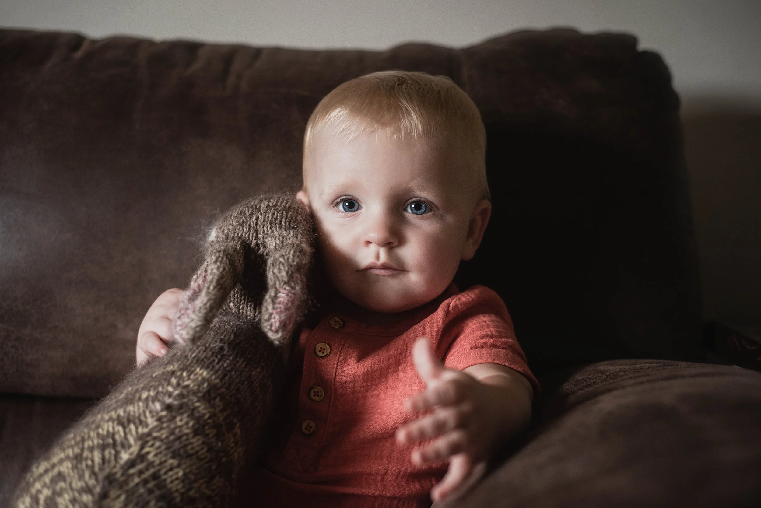 A young child with blond hair and blue eyes sitting on a brown sofa, looking directly at the camera while holding an adult's handwear. The child is wearing a red top with buttons and has a curious expression. Milestone portraits.