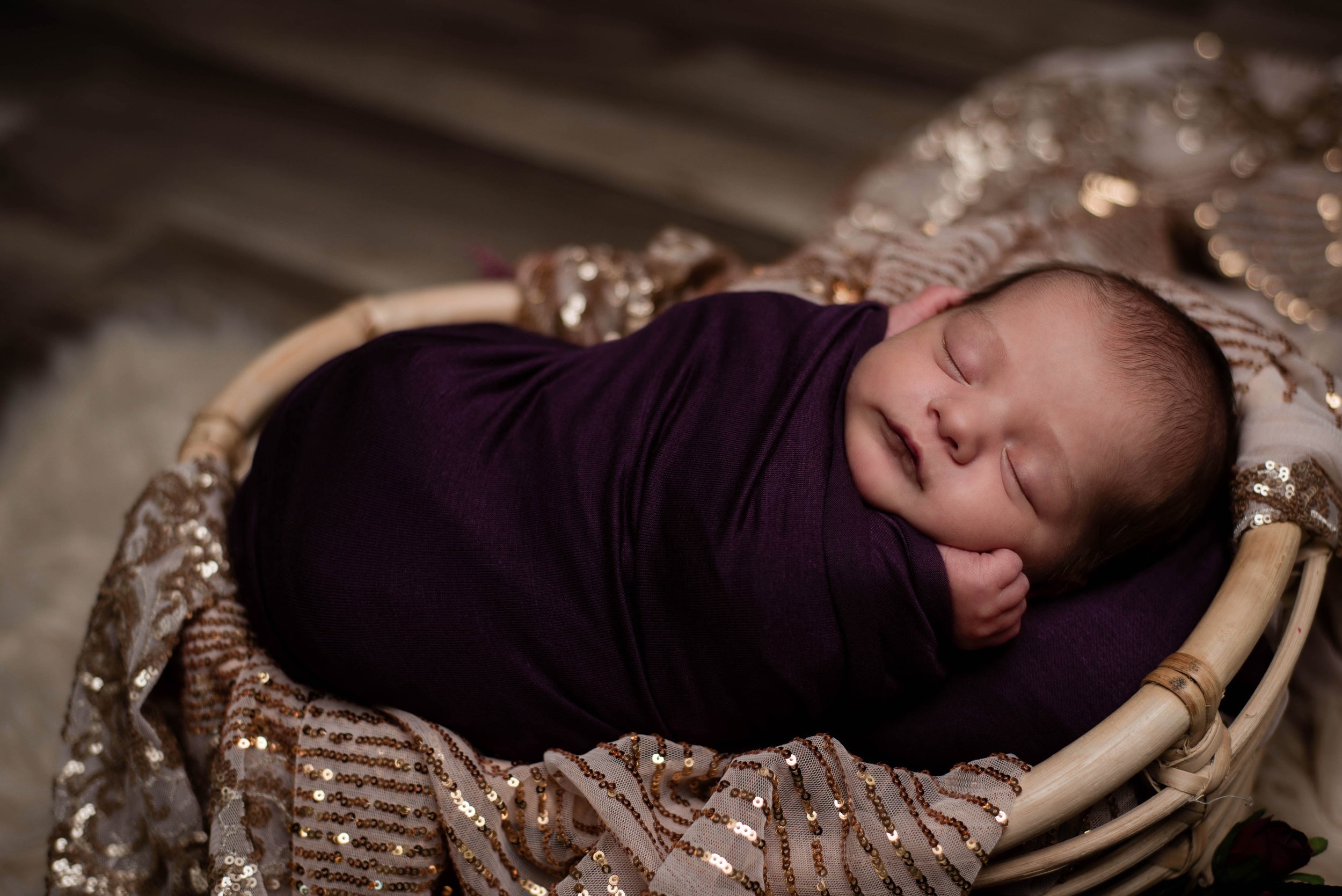 A baby with closed eyes sleeping peacefully in a woven basket, wrapped in a purple cloth, surrounded by a sequined blanket. Studio newborn portrait, Uniontown PA.