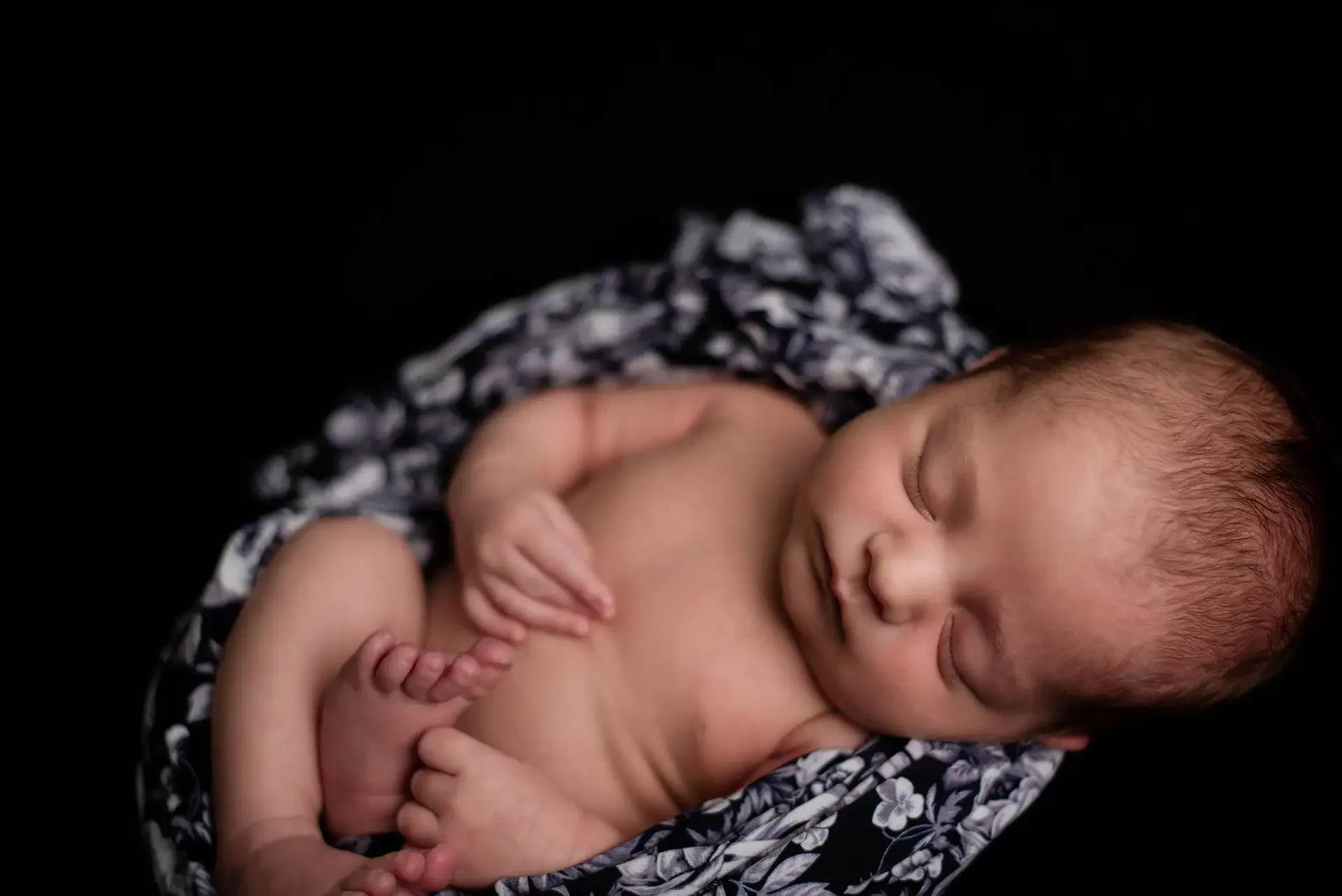 a newborn baby is curled up on a black background, she is wrapped in a black and white swaddle with flowers and skulls, and you can see her hands and feet