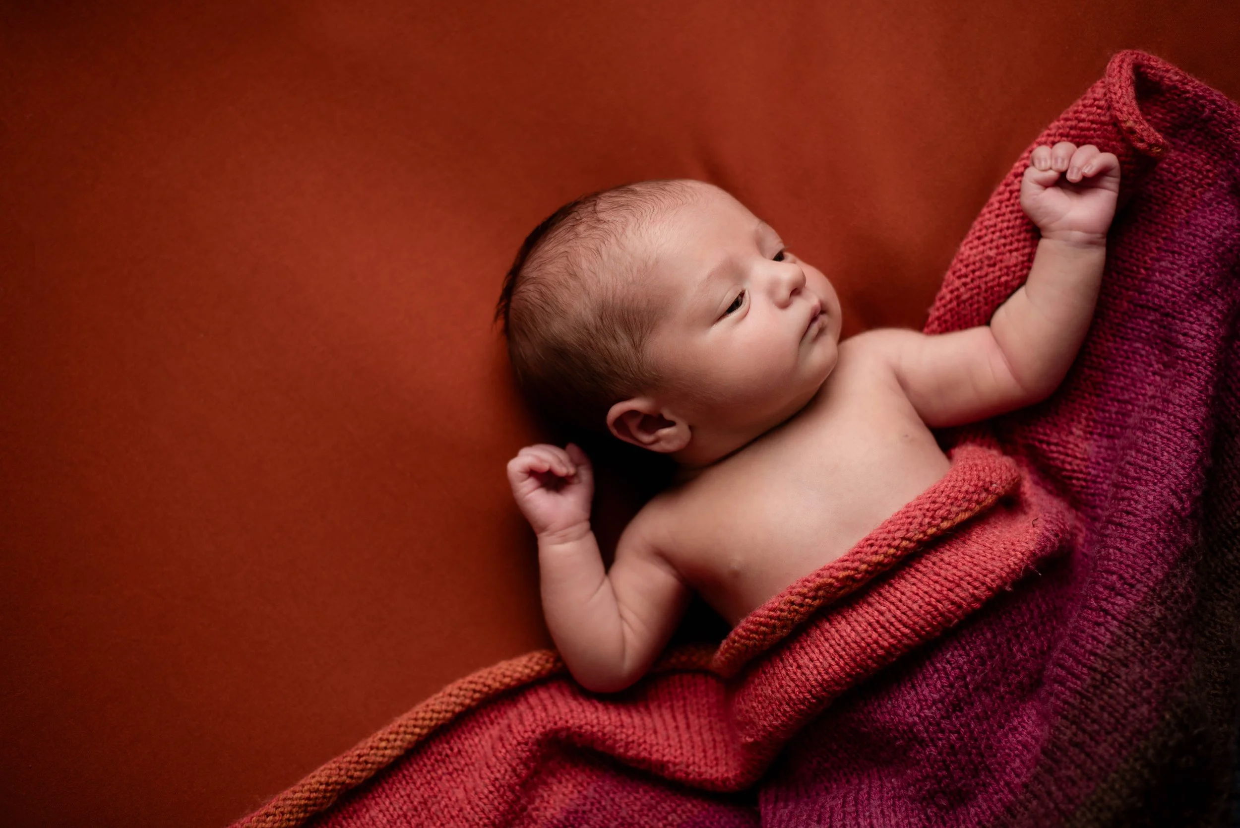 A newborn baby lying on a red blanket and a red background, with arms raised and eyes open. Studio newborn portrait, Brownsville PA.