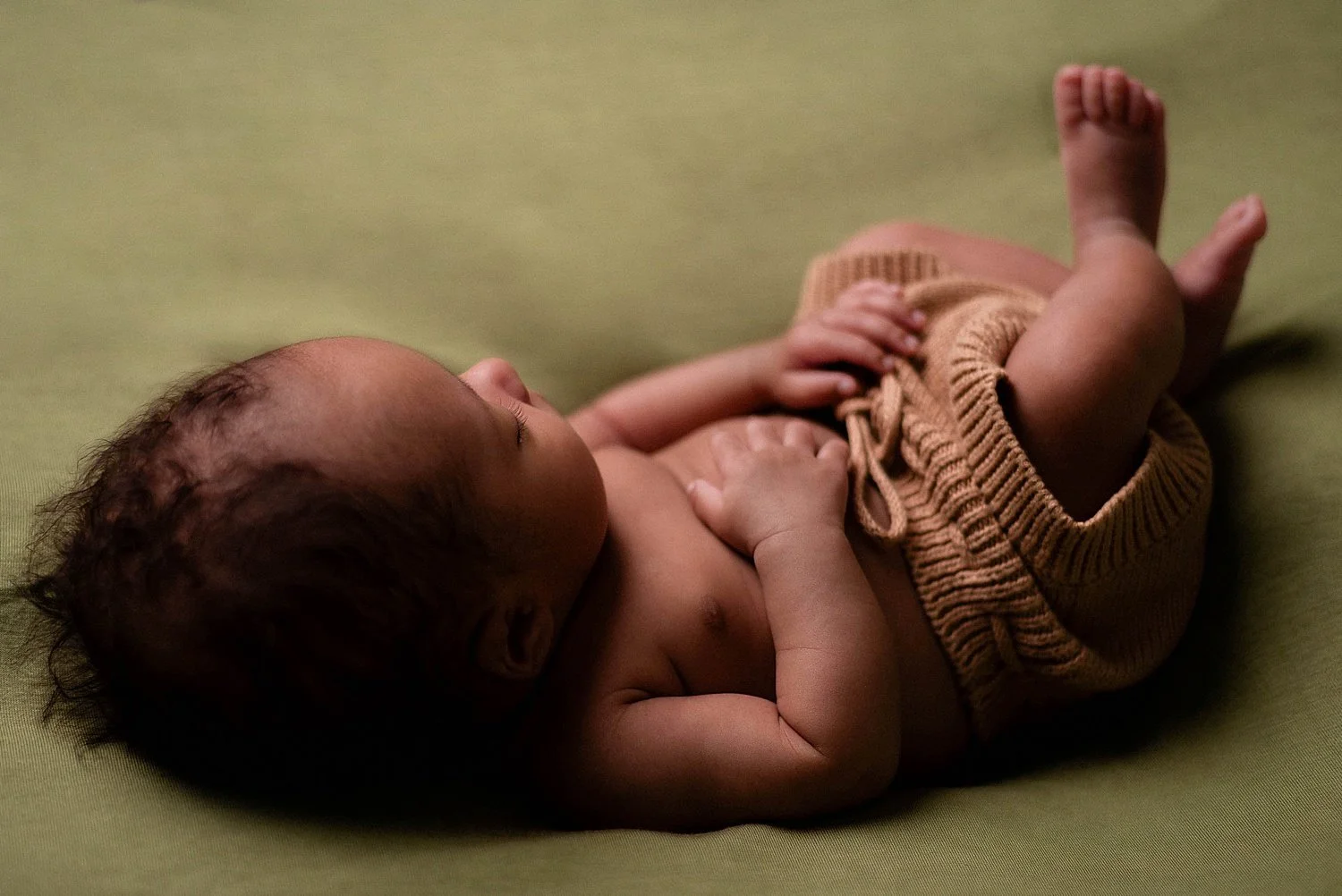newborn baby, black, is wearing knit shorts and resting on a green fabric