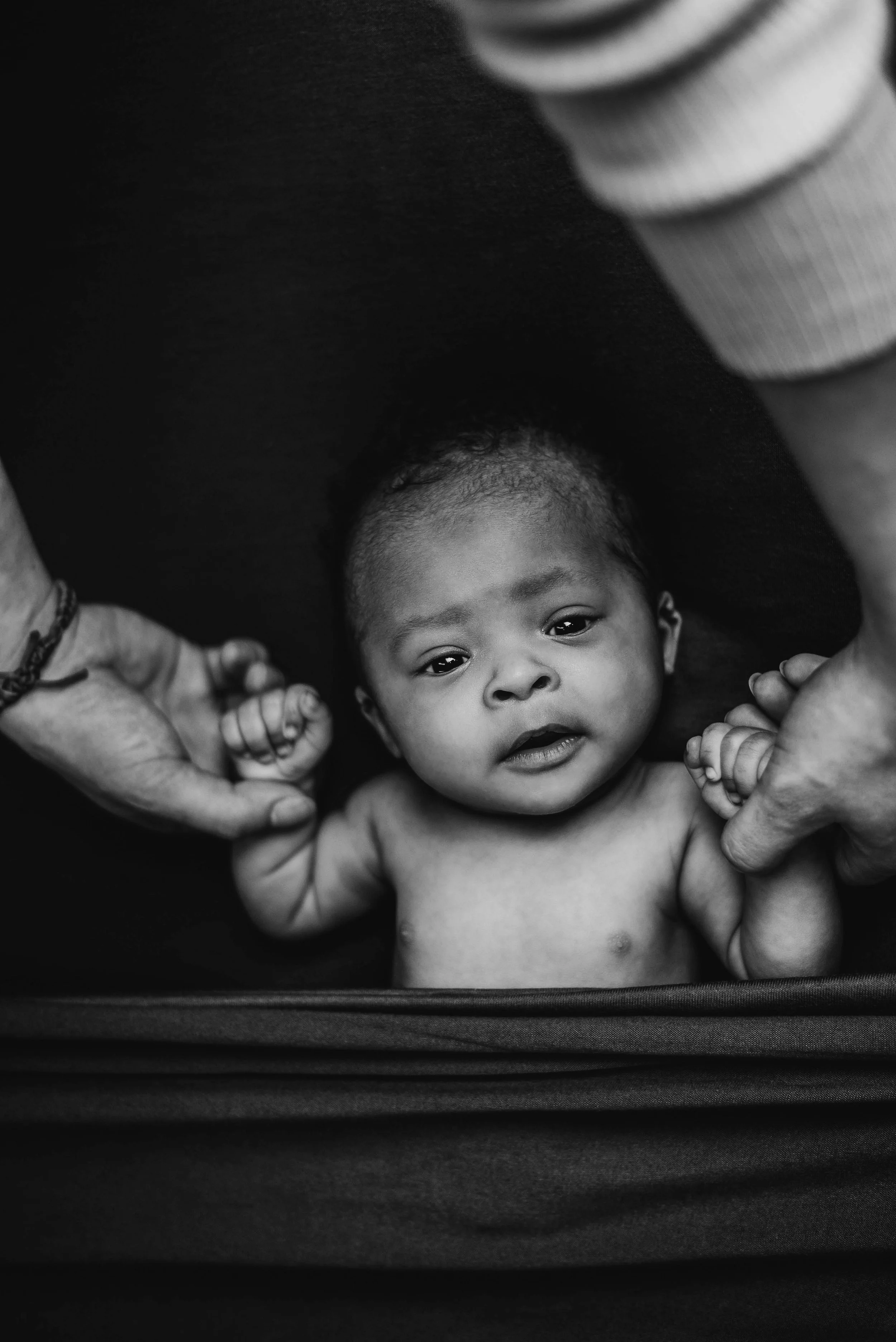 Black and white photo of a baby lying on a mat, held by two adults' hands, with one on each side of the baby, looking up at the camera. Studio newborn portrait, Uniontown PA.