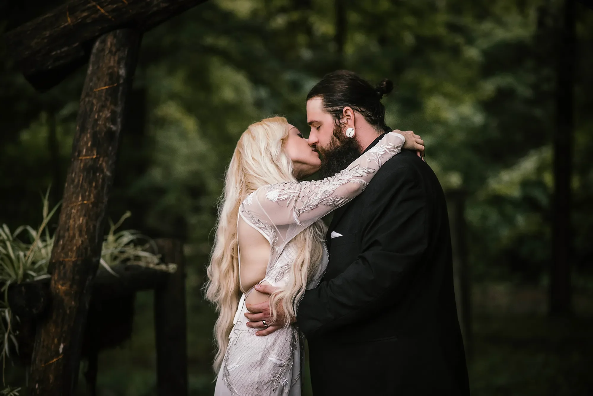 A couple shares a romantic kiss in a forested area, with the woman dressed in a sheer, embroidered dress and the man in a black suit. Elopement at Stonebridge.