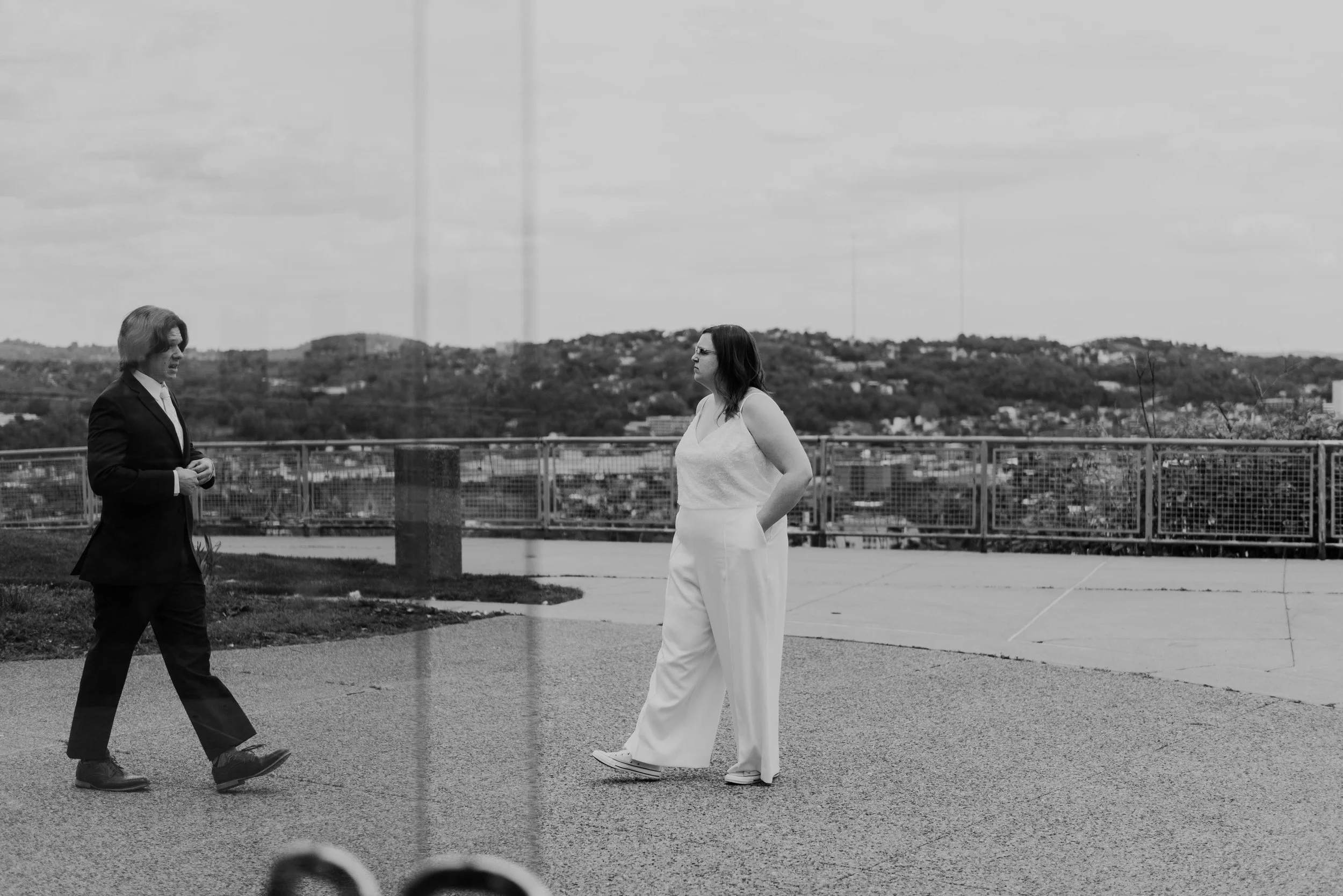 a bride in a white jumpsuit and groom in a dark suit walk toward each other, hands in pockets. behind them is the city of Pittsburgh, at their elopement at West End Overlook