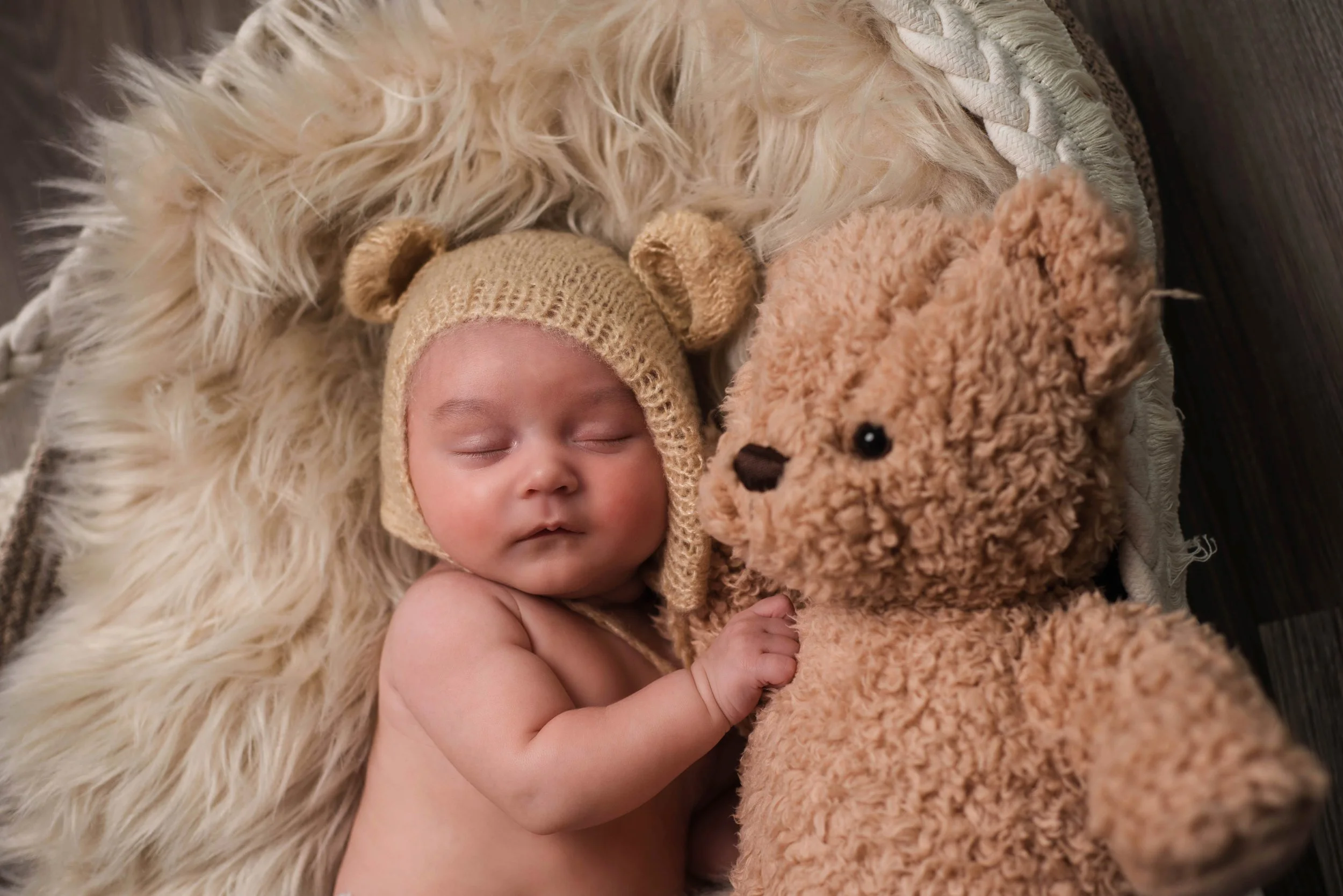 A sleeping baby wearing a beige knit hat with bear ears, lying on a fluffy cream-colored blanket next to a large brown teddy bear. Studio newborn portrait, Brownsville PA.