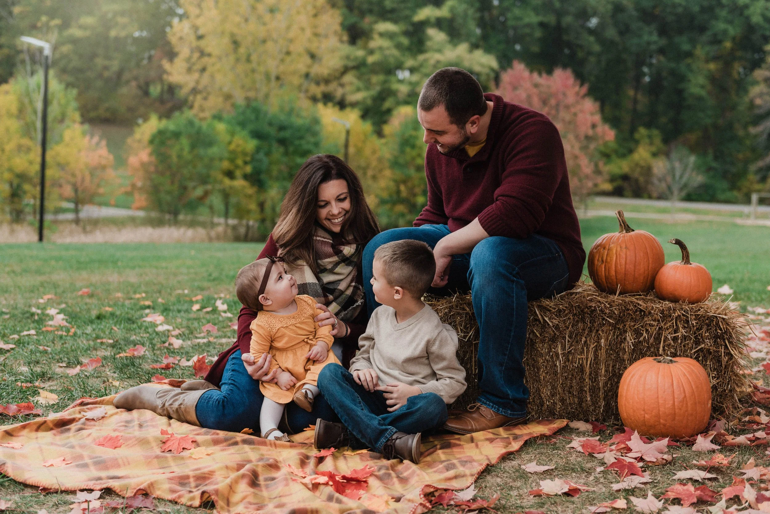 A family of four sits on hay bales and blankets surrounded by pumpkins, they are dressed for fall