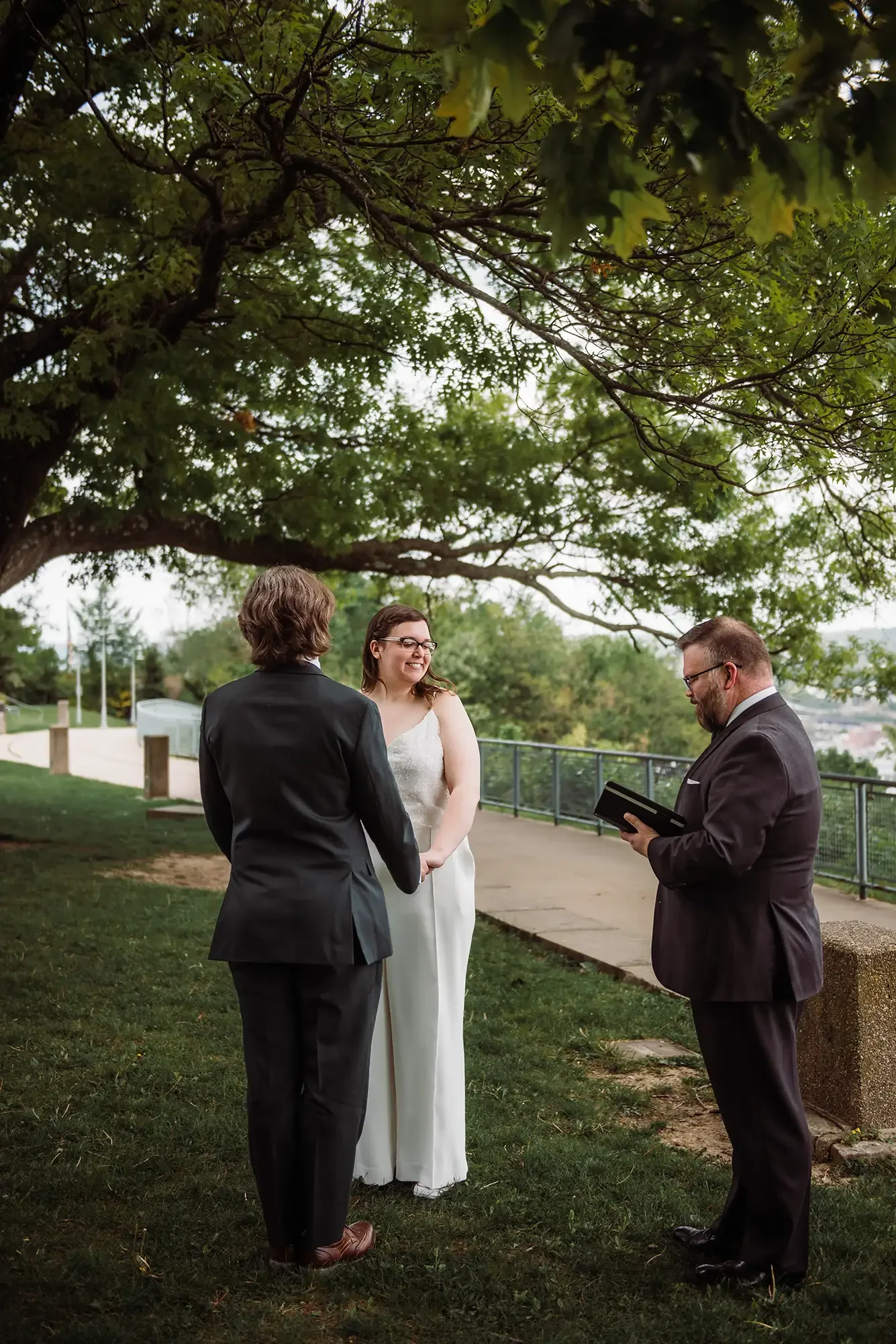 A wedding ceremony taking place outdoors under a large leafy tree, with a man and woman holding hands and facing each other, and an officiant reading from a small book. Pennsylvania elopement in Pittsburgh.