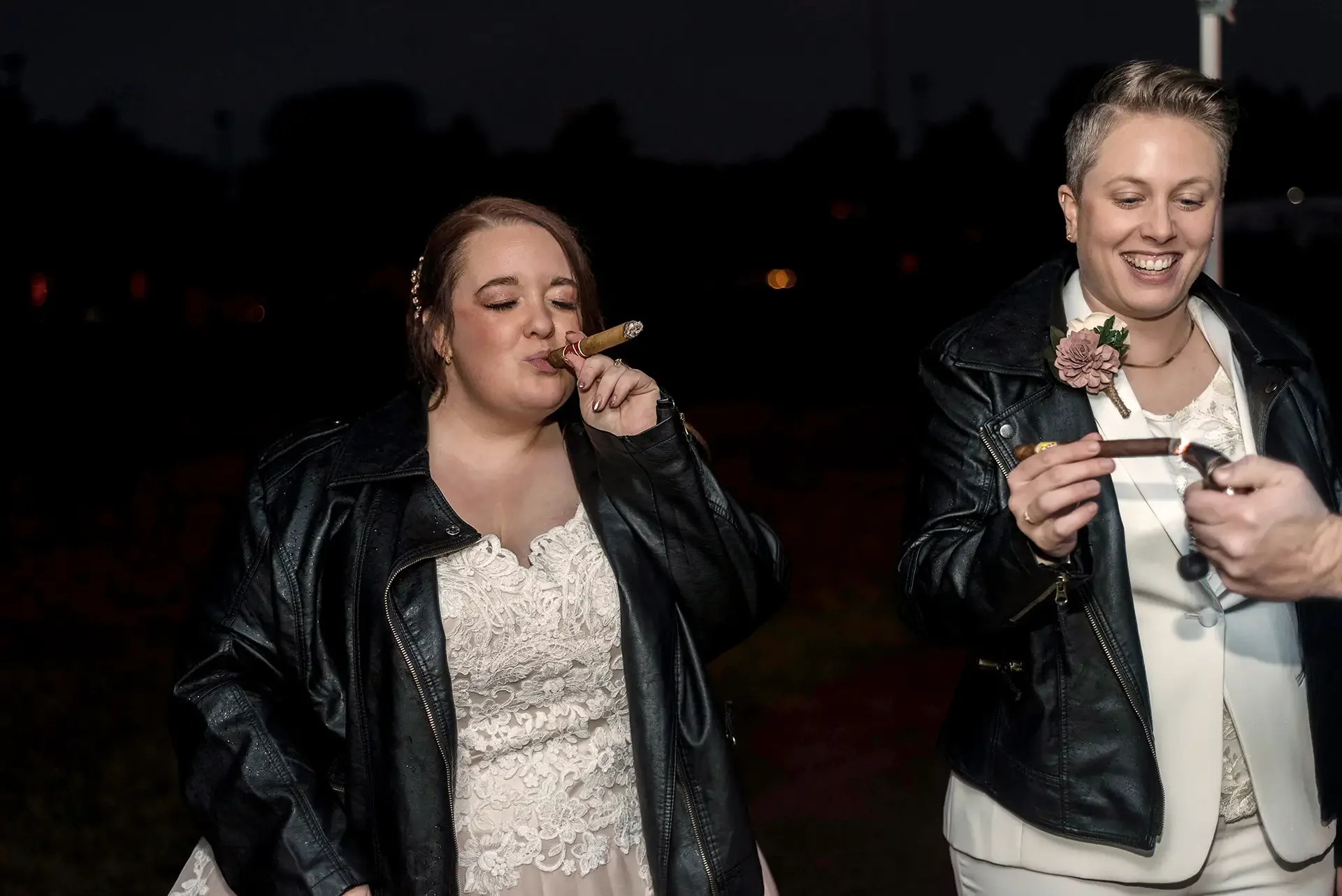 A lesbian couple smoke cigars to celebrate eloping at the Pump House, it is dark and they both wear leather jackets, one over a traditional wedding gown and the other over a cream suit.