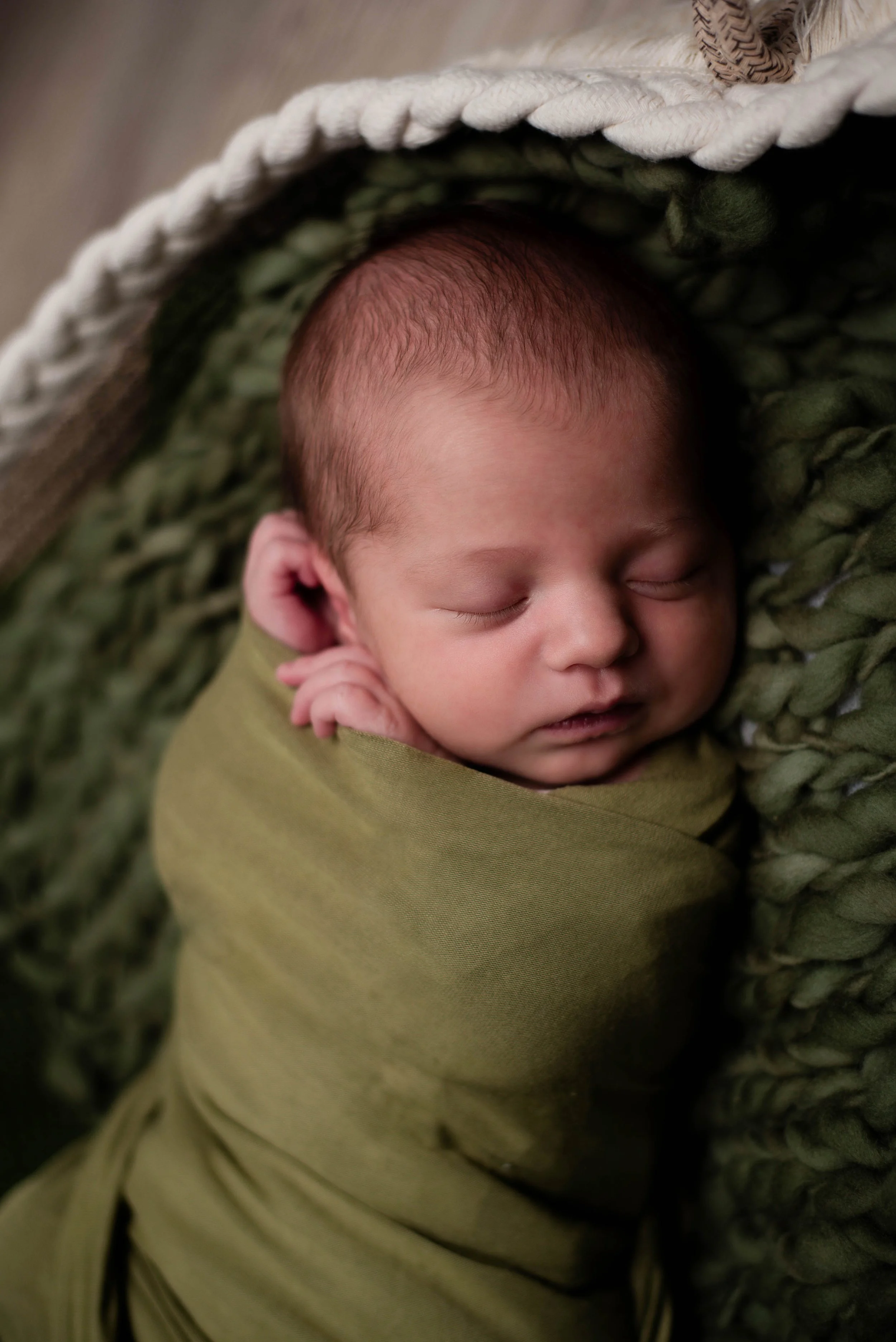 A sleeping baby wrapped in an olive green blanket, lying on a textured green blanket and partially covered by a cream-colored braided blanket. Studio newborn portrait, Brownsville PA.