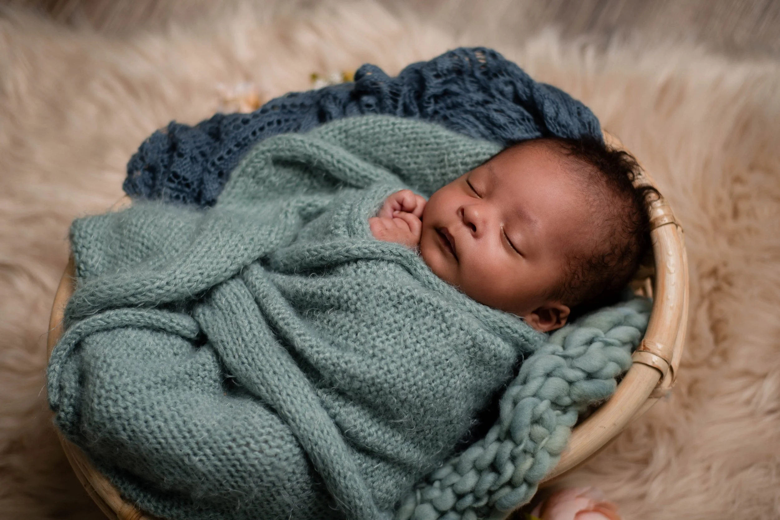 A sleeping baby wrapped in a green knitted blanket in a wicker basket on a soft, fluffy beige rug. Studio newborn portrait, Brownsville PA.