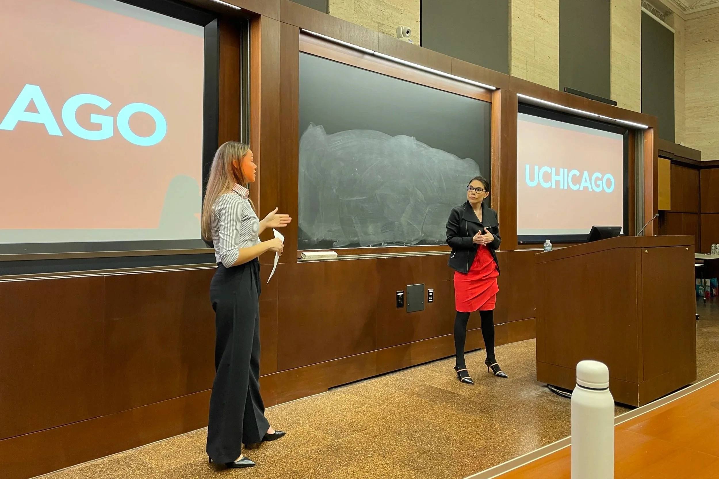 Two women standing at the front of a classroom, one speaking, with a blackboard and two screens displaying the word 'UCHICAGO' in the background. The room has wood paneling and a water bottle on the podium.