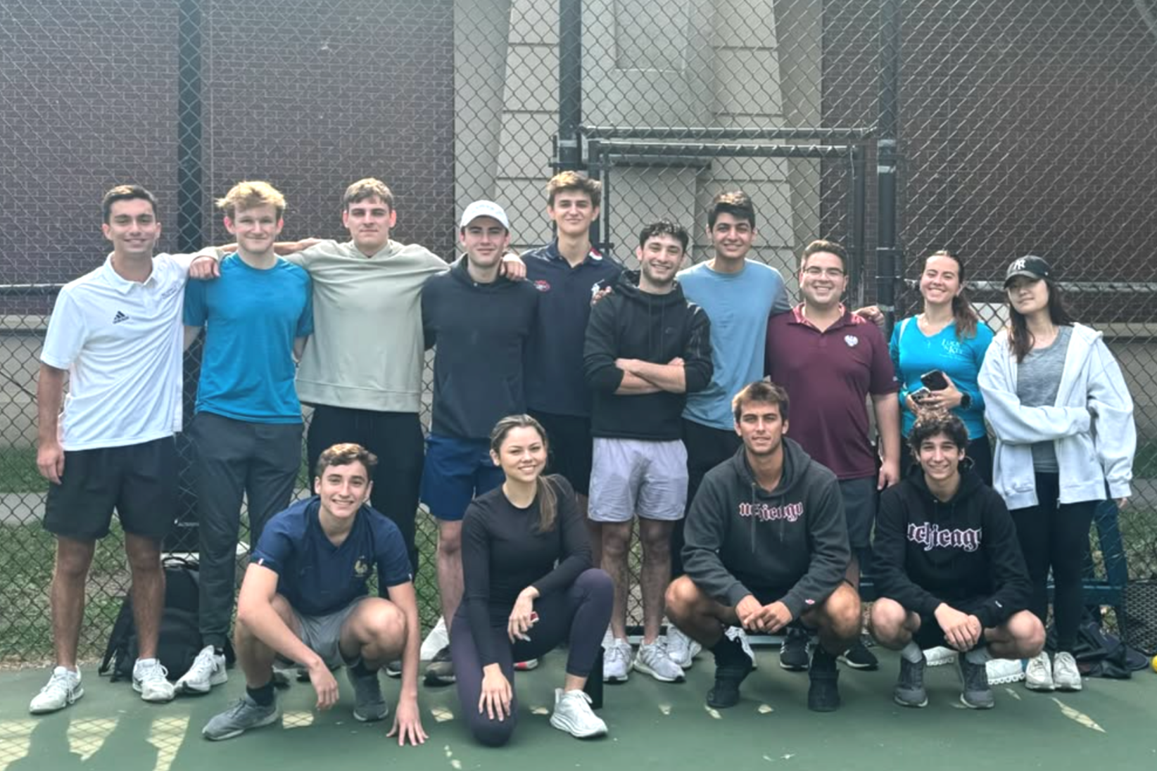 Group of young people on a tennis court, standing and kneeling in front of a chain-link fence, smiling.