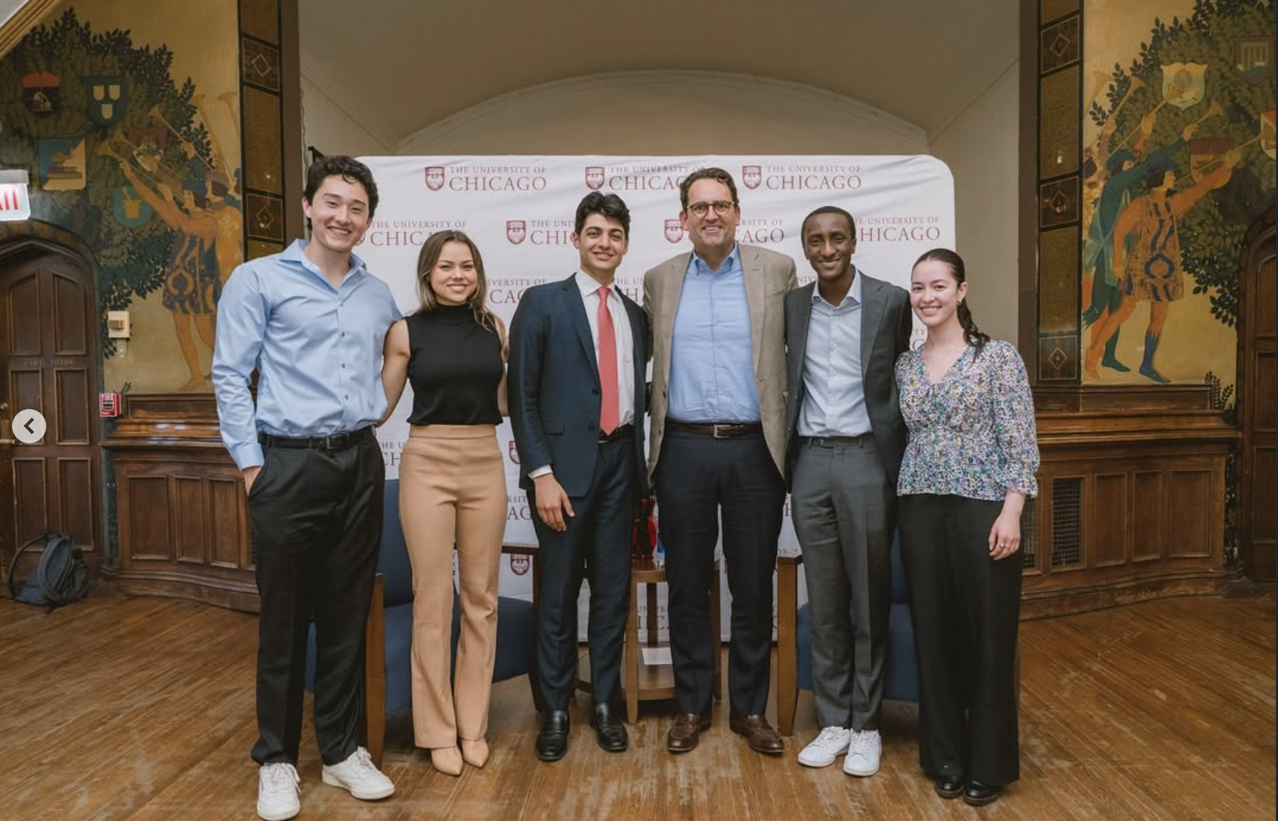 Group of seven diverse young professionals and an older man smiling and posing for a photo at the University of Chicago event, standing in front of a backdrop with the university's logo, on a wood floor in a room with murals on the walls.