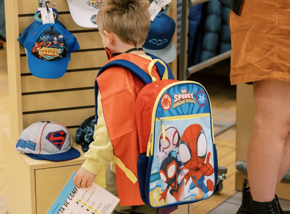 Young boy with a Spider-Man backpack looking at hats in a store. He is holding a paper and wearing a mask. An adult standing nearby is partially visible.
