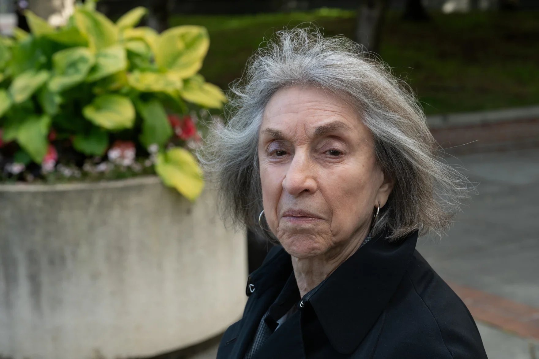 A close-up of an elderly woman with gray, curly hair, wearing hoop earrings and a black jacket, sitting outdoors near a large planter with green and yellow variegated leaves