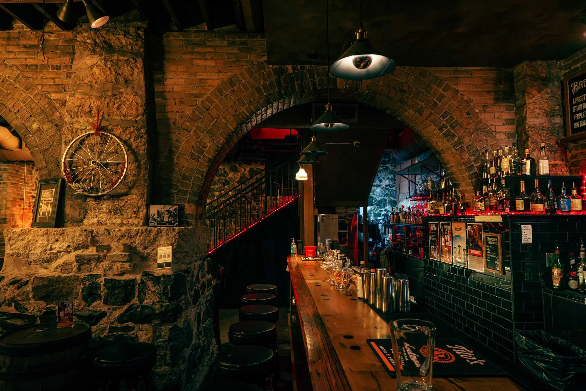 Interior of a bar with brick and stone walls, a wooden bar counter, and a curved brick archway. Shelves behind the bar display various bottles of alcohol, and there are barstools along the counter. Decorative elements include framed pictures and a bicycle wheel mounted on the wall. Lighting is dim with hanging pendant lights.