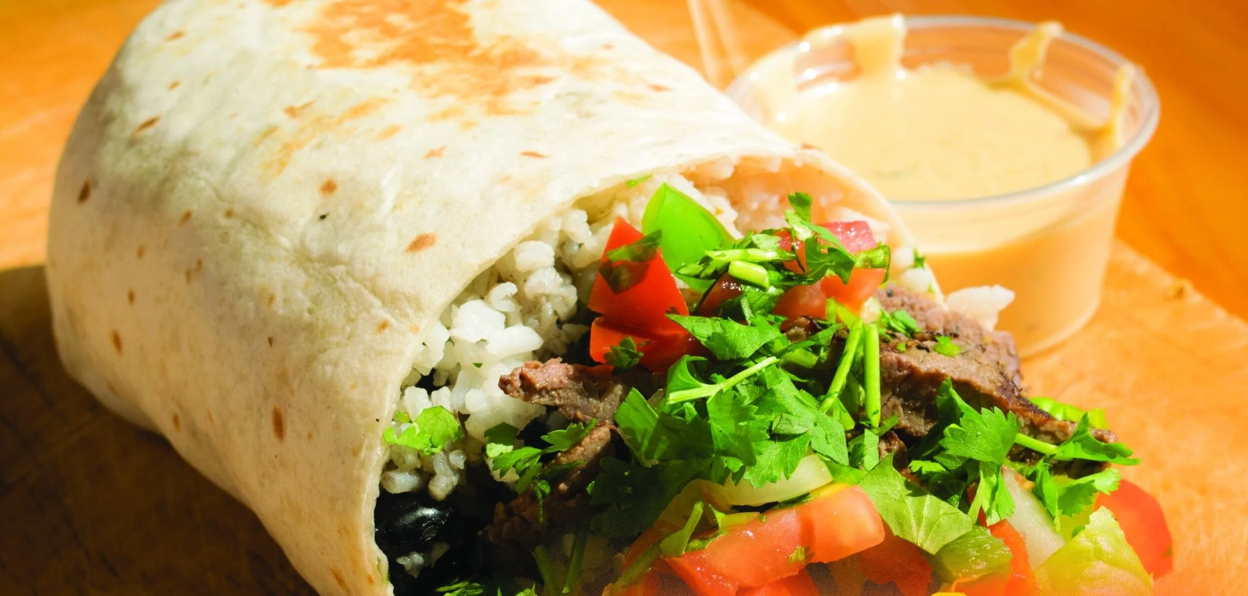 A burrito with rice, ground meat, and vegetables, served with a side of sauce in a small cup on a wooden surface.