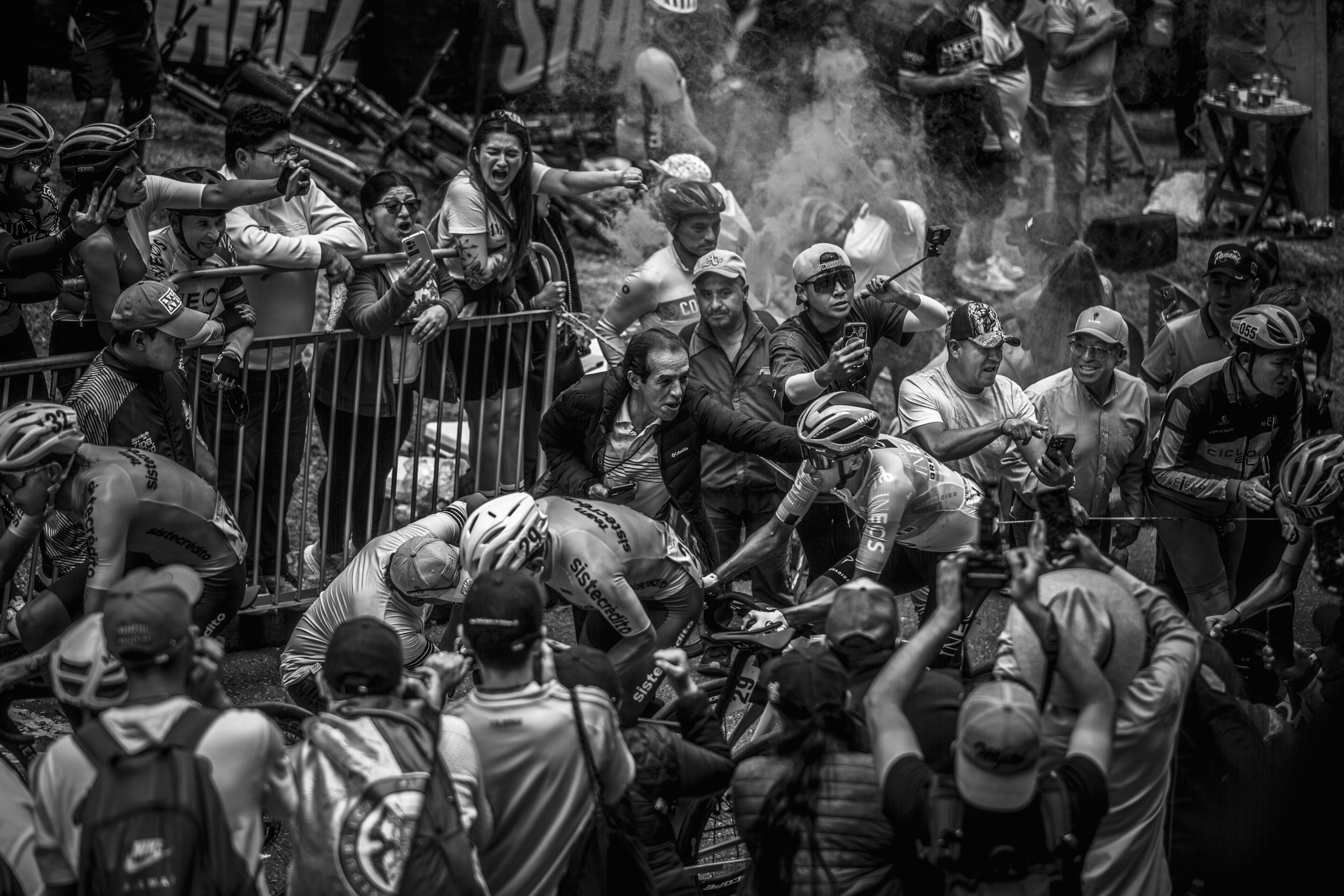 Varios ciclistas en una competencia, con espectadores y fotógrafos alrededor en un ambiente intenso y competitivo.