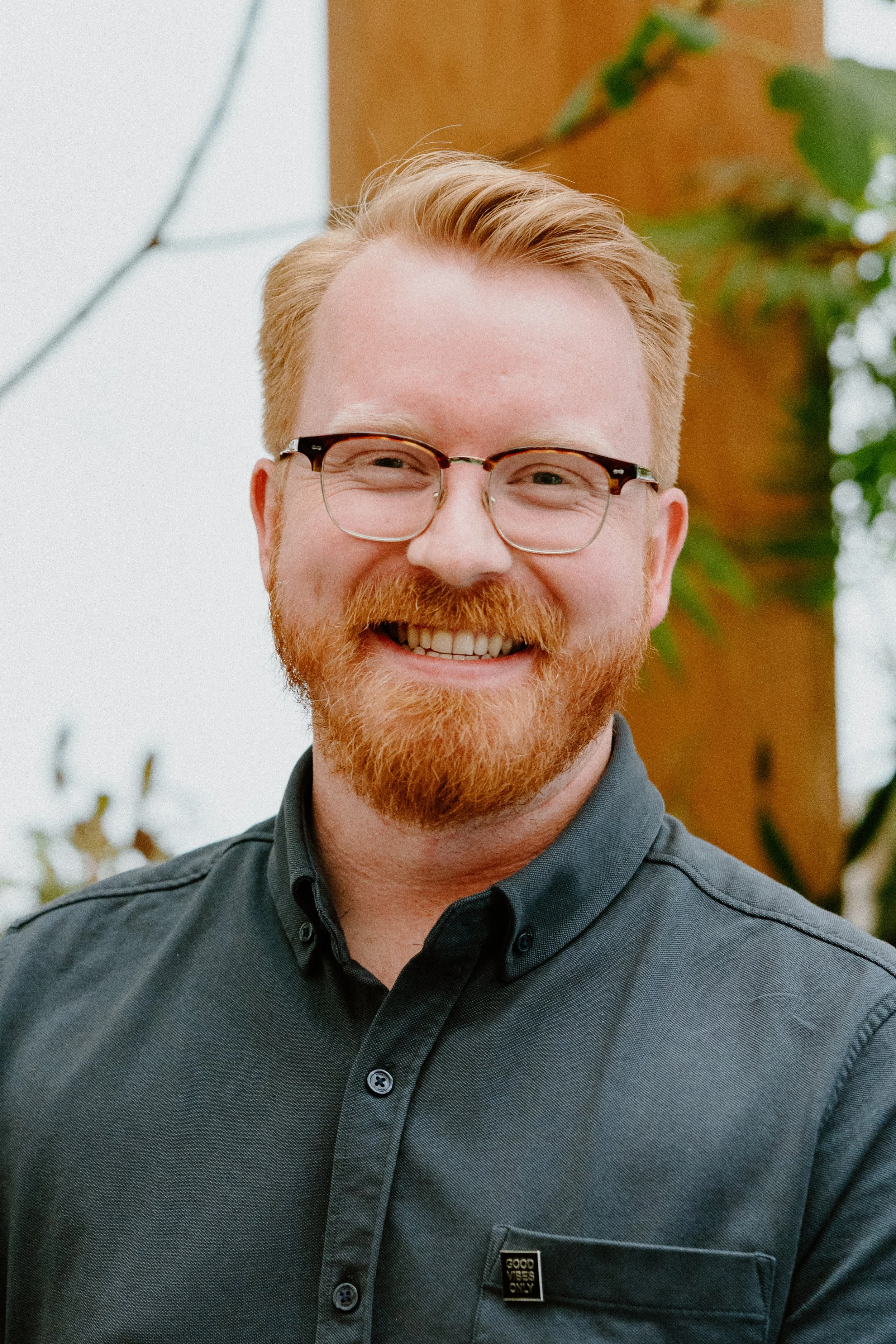 Smiling man with red hair and beard, wearing glasses and a dark gray button-up shirt with a small pin that says 'Good Vibes Only'.
