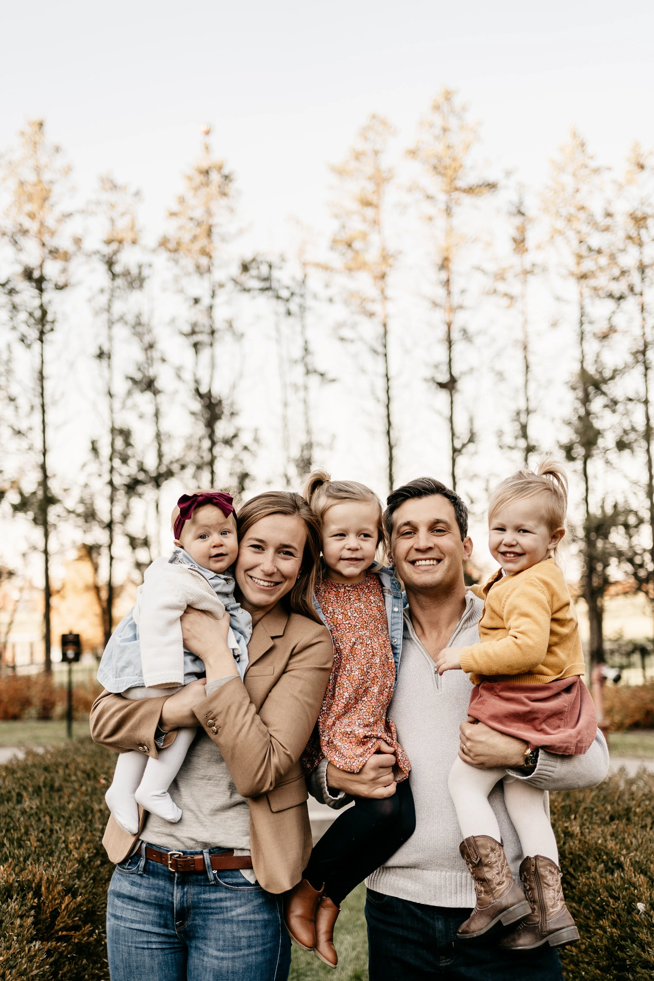 family of father, mother, and three young daughters, standing together with heads close, all smiling, outside in front of trees