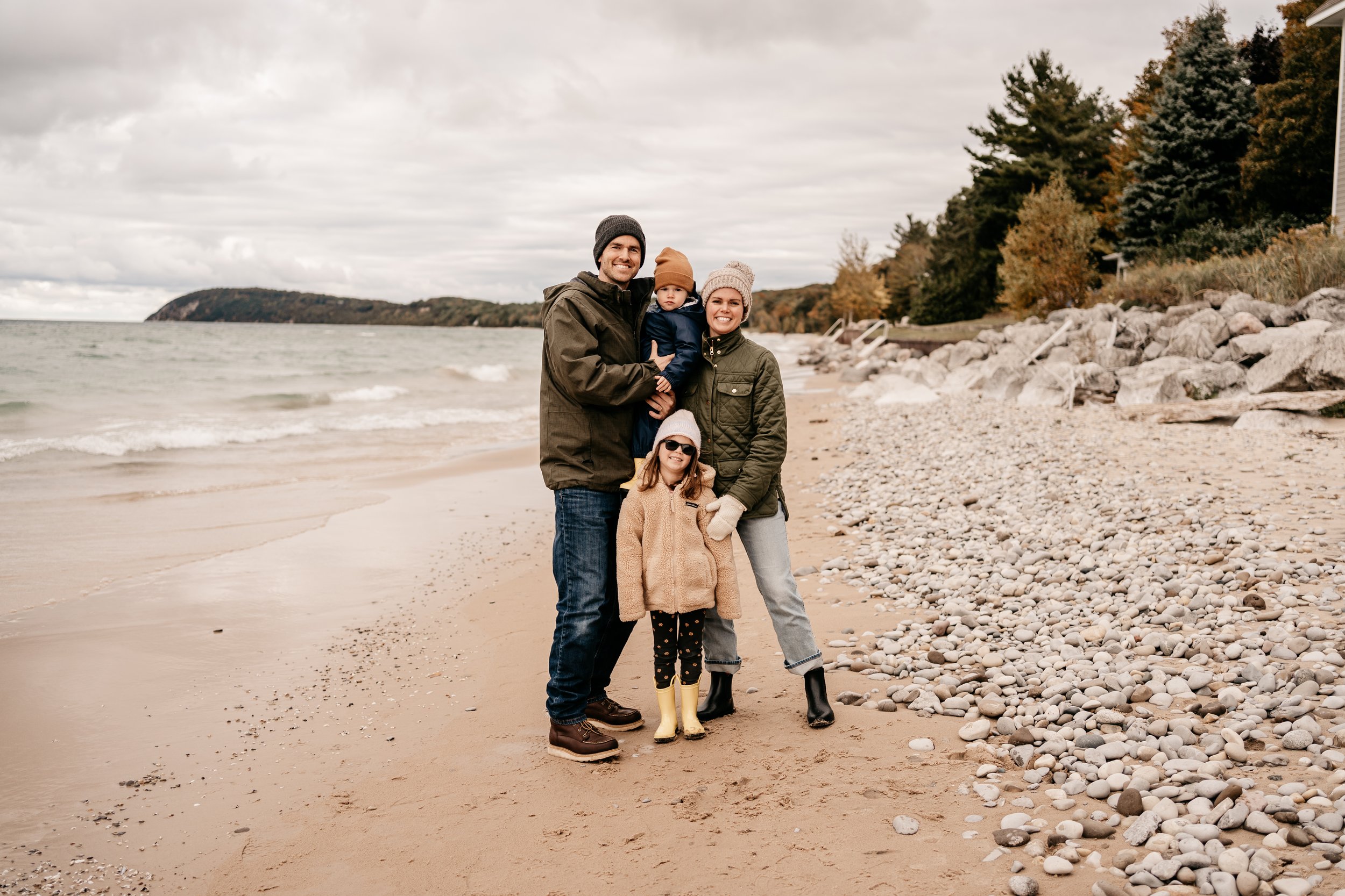 family of four - father, mother with no hair due to breast cancer, young daughter, and toddler son, standing together and smiling on the shore of lake Michigan in Leland