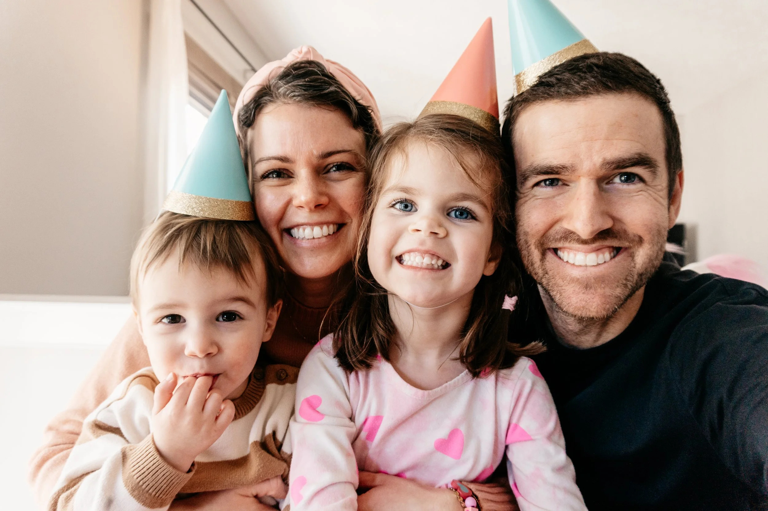 young mother of two diagnosed with breast cancer in selfie with husband, young daughter, and toddler son, wearing birthday hats