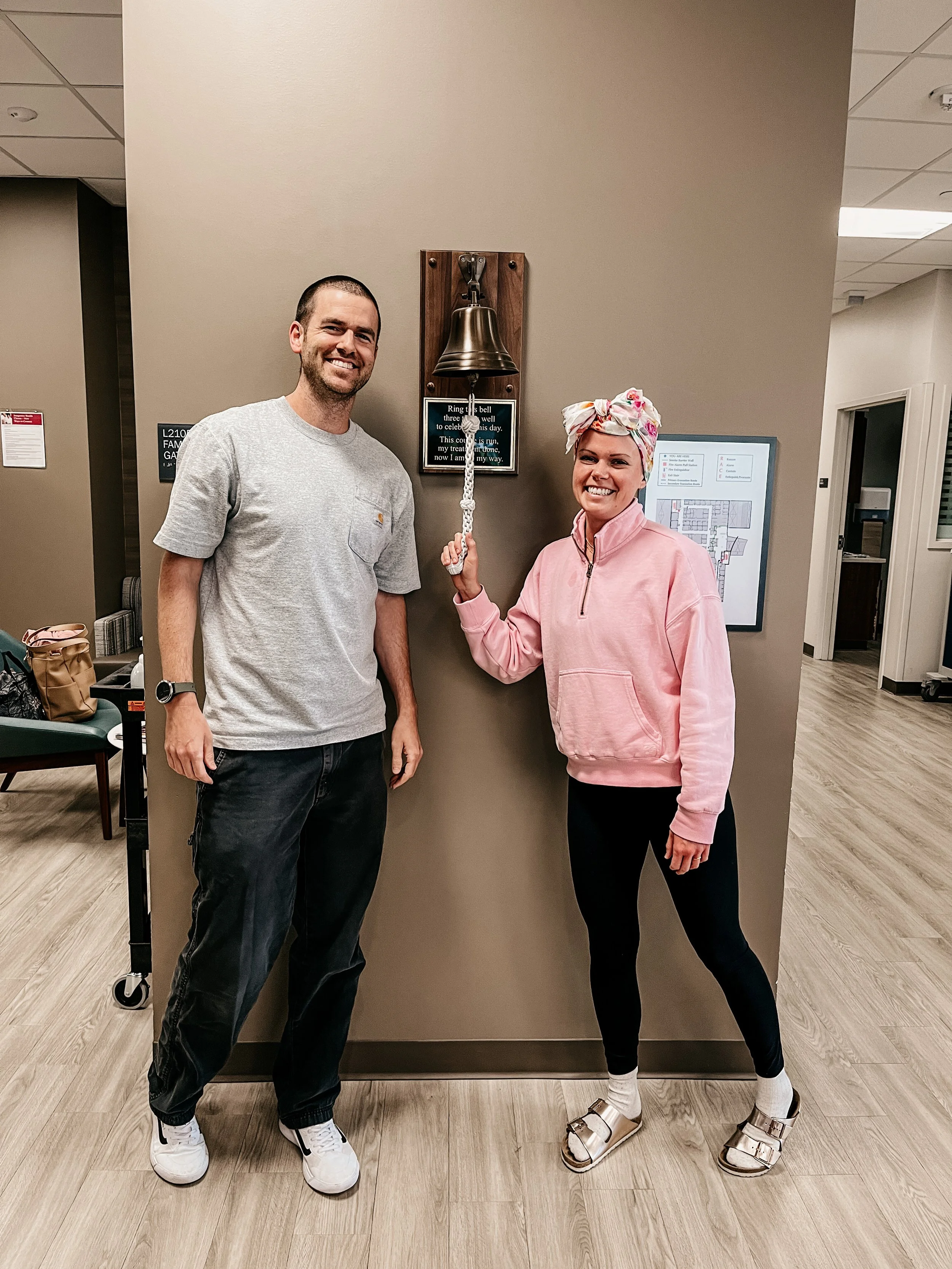young mother of two diagnosed with breast cancer ringing bell after completing chemo treatment. wearing black leggings, pink sweatshirt, and floral head scarf. Husband stands next to her.