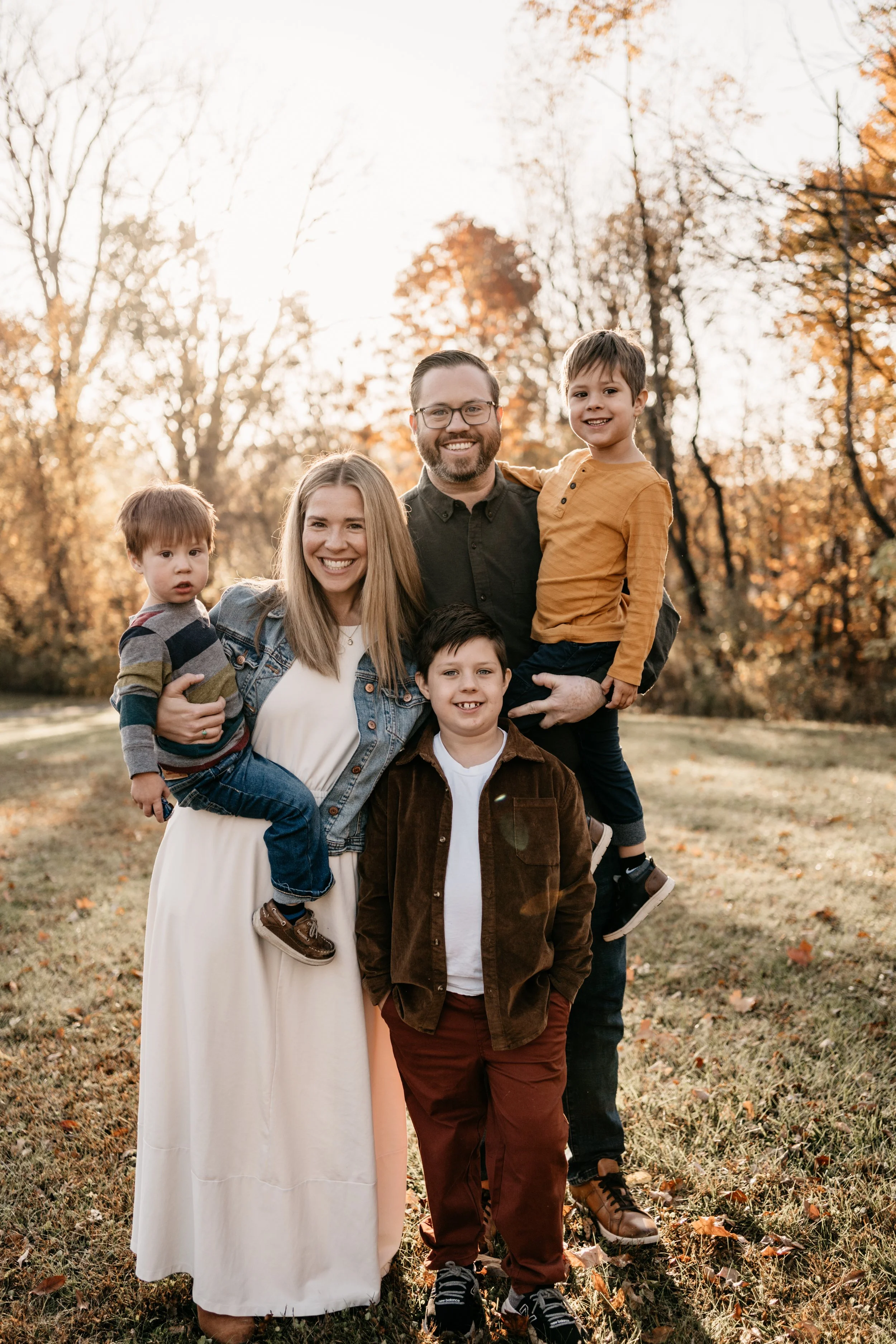 family of mother, father, and three young sons, standing together and smiling, in front of trees on a fall day
