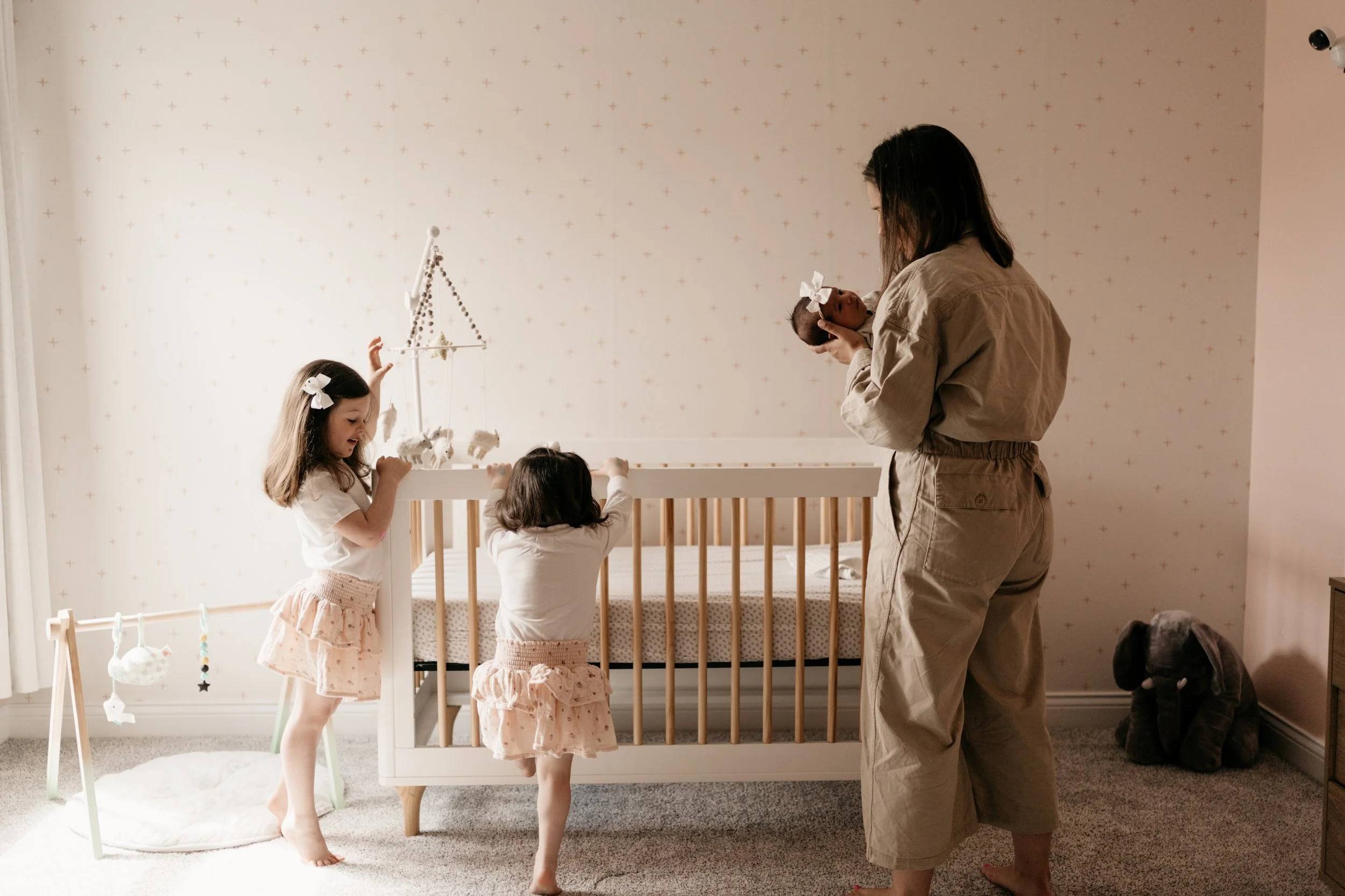 Mother holding baby by crib, two younger sisters standing next to crib
