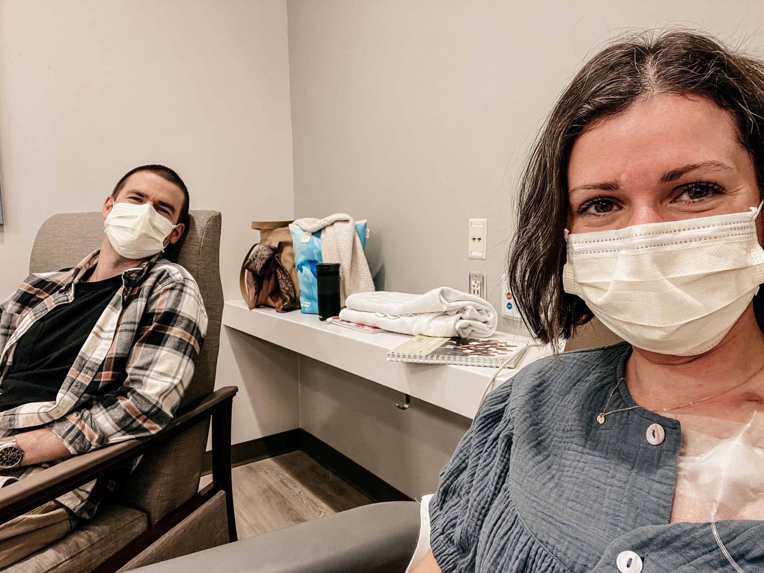 selfie of woman with mask on in chemo chair during treatment, with husband in chair next to her