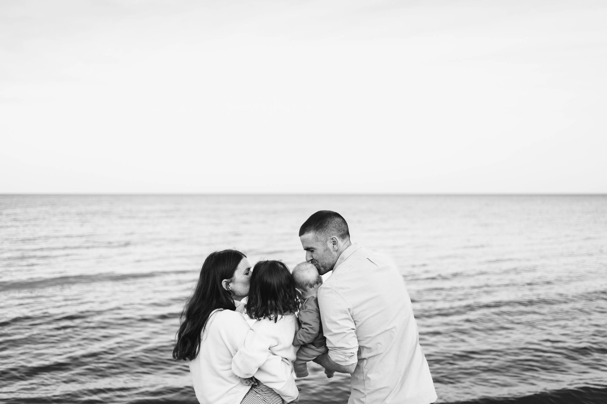black and white photo of mom, dad, young daughter, and baby son. all facing the water, parents are kissing the kids that are in the middle.