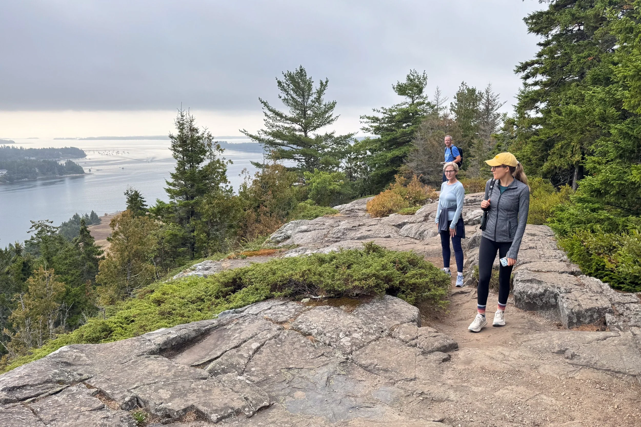 Three people hiking on a rocky trail in a forested area overlooking a body of water and an overcast sky.
